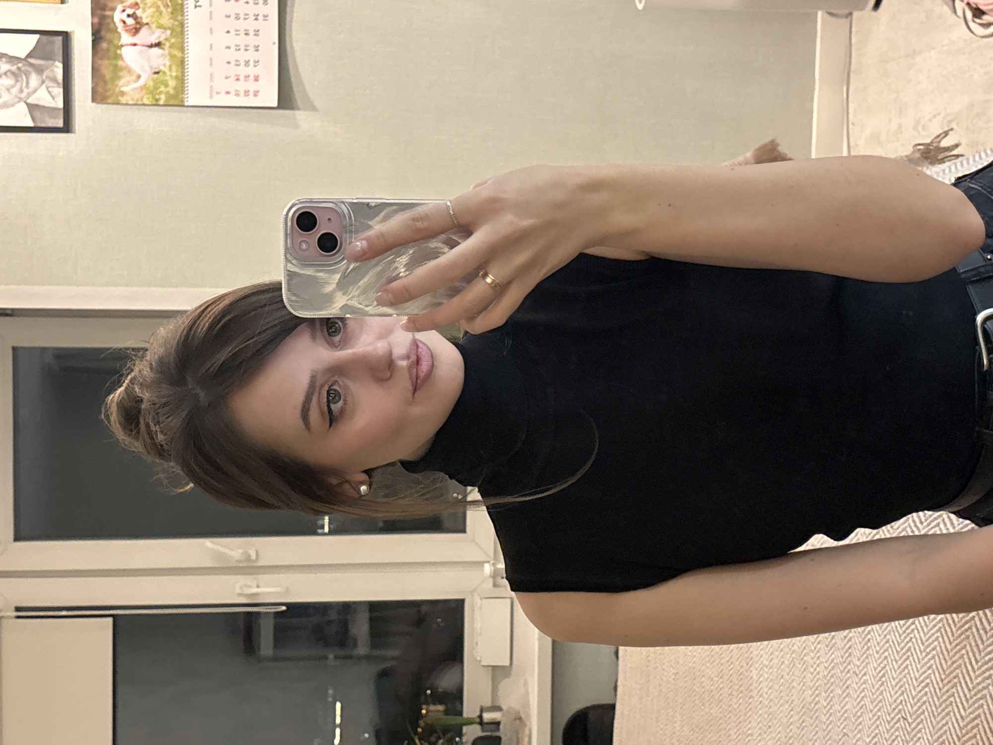 The image depicts a female individual, approximately 20-35 years of age, taking a mirror selfie indoors at night in Krasnoyarsk, Russia. The individual has medium-brown hair styled in an updo with bangs, light-colored eyes with winged eyeliner, and wears small pearl-like stud earrings. She is dressed in a black sleeveless turtleneck top and has a thin gold-colored ring on her left ring finger and a thin silver-colored ring on her left pinky finger. She holds a light pink smartphone, encased in a clear or iridescent case with a three-lens camera module, in her left hand.
The background shows a room with light-colored walls. To the left, a large white-framed window reveals a dark exterior with distant city lights. A white windowsill or shelf below the window holds a dark potted object and a small white bottle. On the wall above the individual's right shoulder, a framed black-and-white portrait of an older man hangs above a wall calendar. The calendar displays an image of a small, light-colored dog defecating on grass and features text primarily in Cyrillic script. A patterned fabric, possibly from a bed or couch, is visible in the lower left, and a light-colored floor is visible in the lower right.