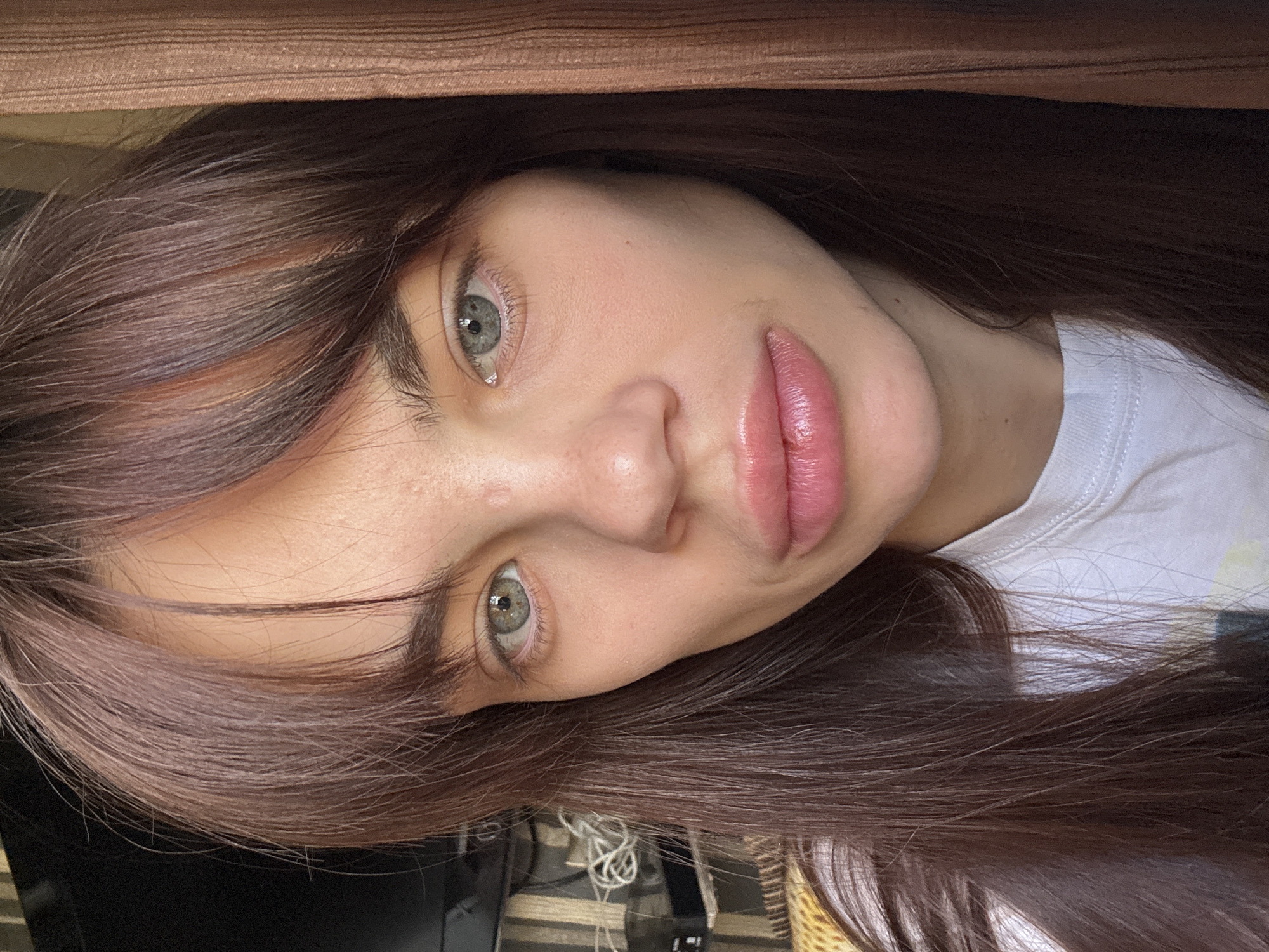 A young woman with long brown hair and green eyes is shown in a close-up, slightly tilted portrait. She has fair skin and pink lips. She is wearing a white shirt. A brown object is visible in the upper part of the frame, above her head. The image is taken indoors. There is no indication of Rostov-on-Don, Russia, or any specific city.