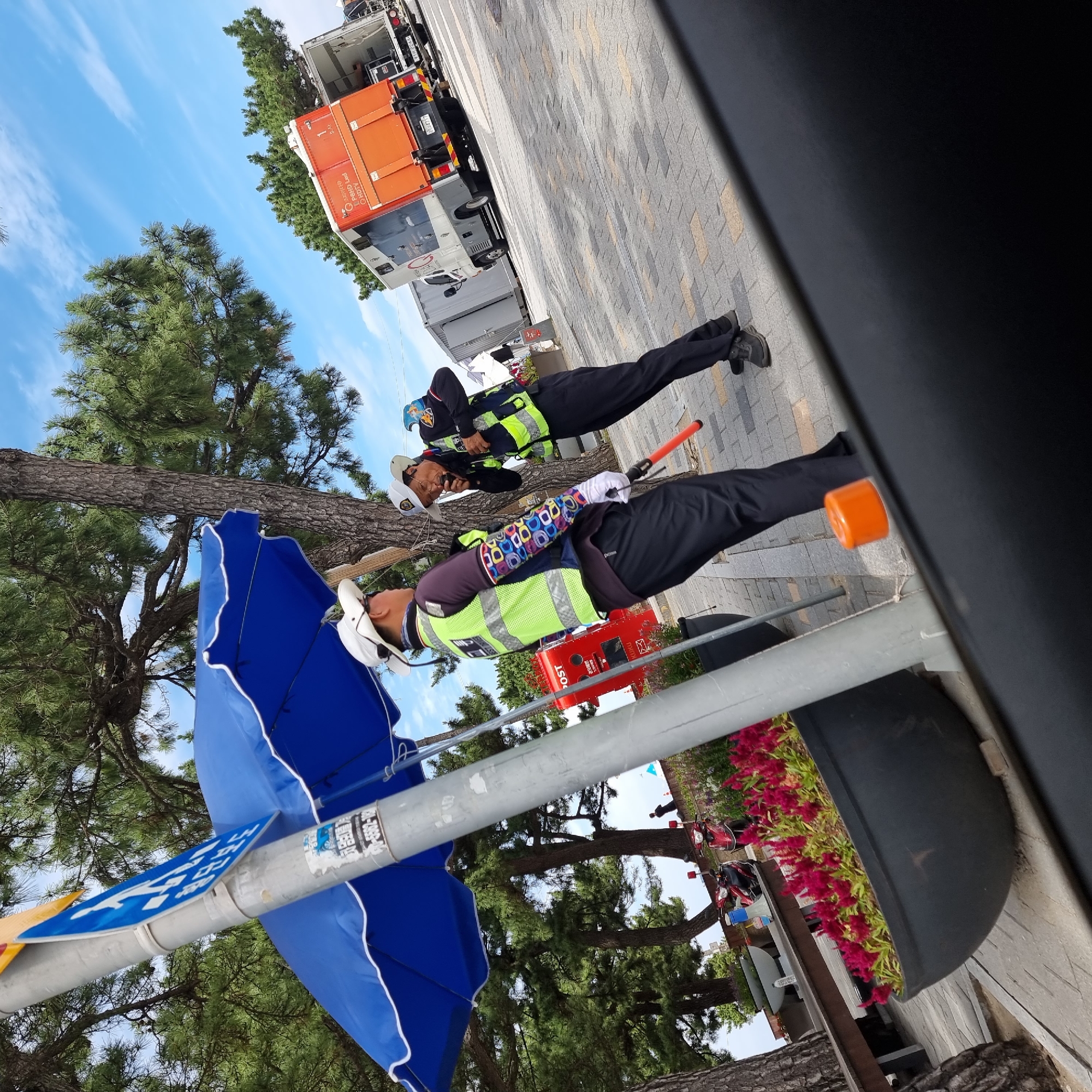 In Gangneung-si, South Korea, two individuals wearing high-visibility vests and white hats are interacting near a pole with a blue umbrella. One individual is facing left, holding an orange signaling stick, and appears to be speaking. The other individual is facing right and speaking into a radio. Behind them, a bright orange truck with a white trailer is parked, and to the left, a red postal box is visible. The ground is paved with a brick pattern. Pine trees and a bright blue sky with some clouds form the background. Red flowers are planted in a large black pot in the foreground.
