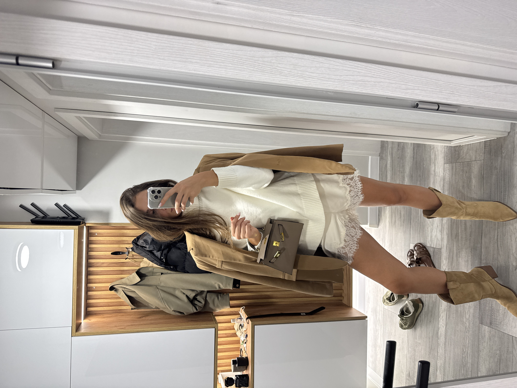 A young woman stands in a hallway, taking a mirror selfie. She is wearing a white turtleneck sweater, white lace-trimmed shorts, a tan blazer draped over her shoulders, and tan suede cowboy boots. She holds a small brown handbag in her left hand. The hallway features white walls and white doors, with a wooden accent wall in the background where several coats are hanging. On the floor, to the left of the woman, are a pair of green sneakers and a pair of brown sandals. The location is identified as a hallway in Burgas, Bulgaria.