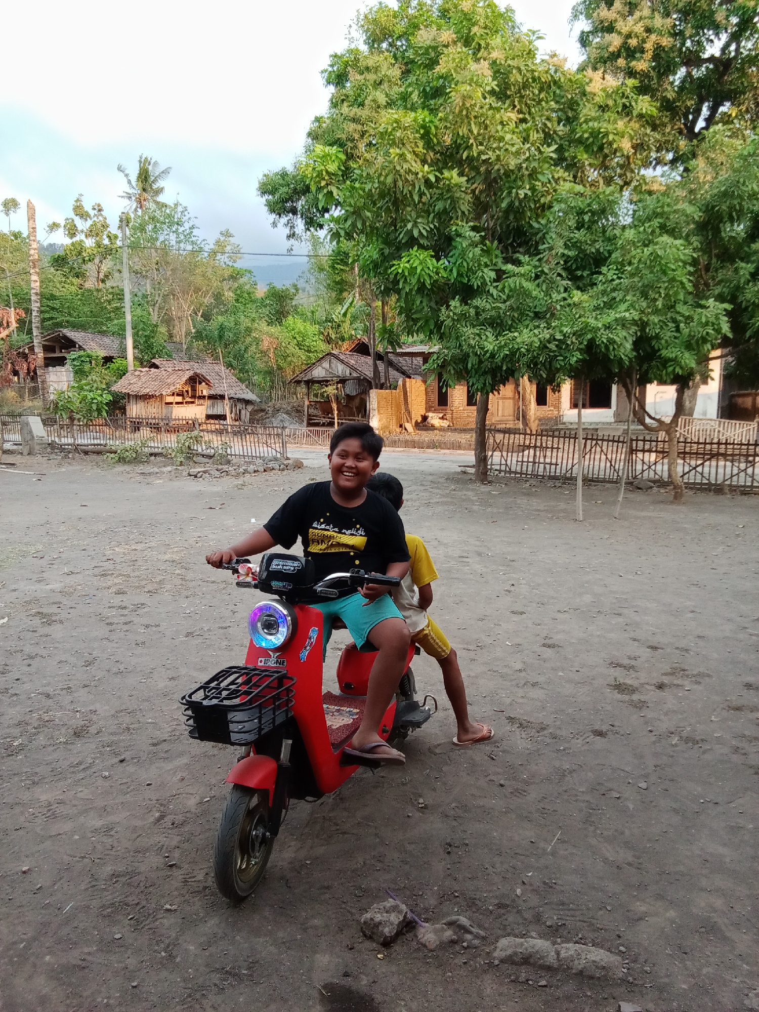 Two young boys are on a red scooter in a rural area, possibly in Selowogo, Indonesia. The boy in the front is wearing a black t-shirt and blue shorts, smiling at the camera. The second boy, wearing a yellow t-shirt and yellow shorts, is seated behind him. They are on a dirt road, with a small village and lush green trees in the background. The scene appears to be set in the late afternoon, with warm lighting.