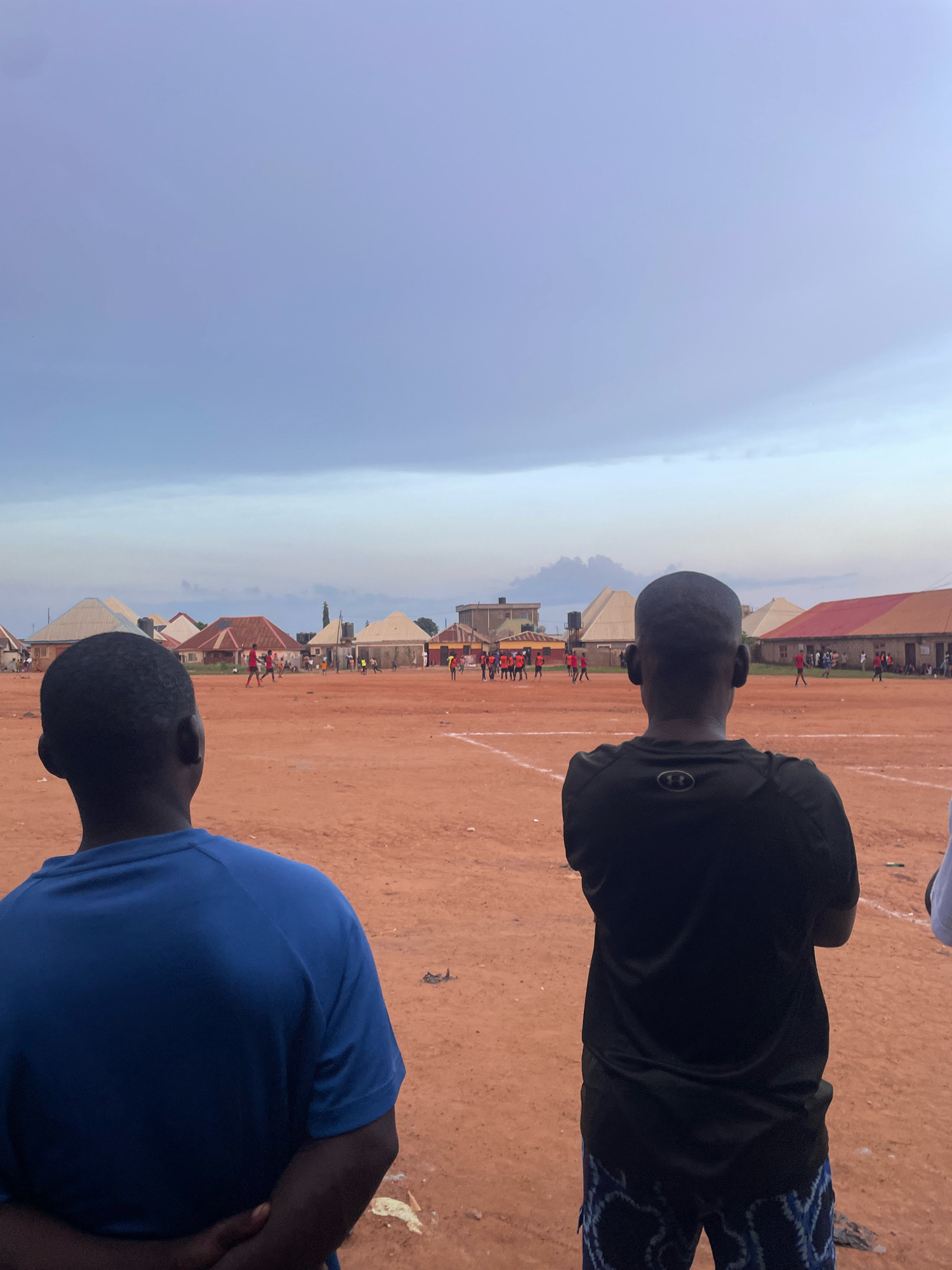 The image depicts an outdoor scene in Kurunduma, Nigeria, possibly a sports field. Two individuals are positioned in the foreground, facing away from the camera and towards a distant activity. One person, on the left, wears a blue t-shirt and has their arms crossed behind their back. The other person, on the right, wears a black t-shirt with a logo on the back and patterned shorts.
In the middle ground, a group of people, some wearing red shirts, appear to be engaged in a football match on a reddish-brown dirt field. The field is marked with white lines. Several buildings with distinctive sloped roofs are visible in the background, suggesting a residential or community area. The sky above is overcast with muted colors, hinting at either dawn, dusk, or a cloudy day. Scattered debris is present on the ground in the foreground and middle ground.