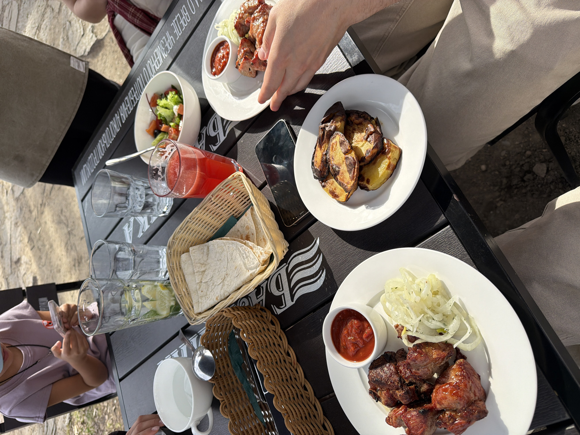 A top-down shot shows a dining table in an outdoor setting, likely in Krasnoyarsk, Russia, based on the Cyrillic text on the table. The table is set with various dishes: two plates of grilled meat with sauce, one with raw onions and the other with what looks like lettuce, a plate of fried potatoes, a bowl of salad, a basket of flatbread, two glasses of juice, and several empty glasses. A smartphone lies on the table next to the food. Two people are partially visible, one wearing beige trousers and reaching for the grilled meat, and another wearing a mask and a pink shirt, holding a small white object. The table is black with gray Cyrillic writing on the side and is placed on an outdoor terrace.