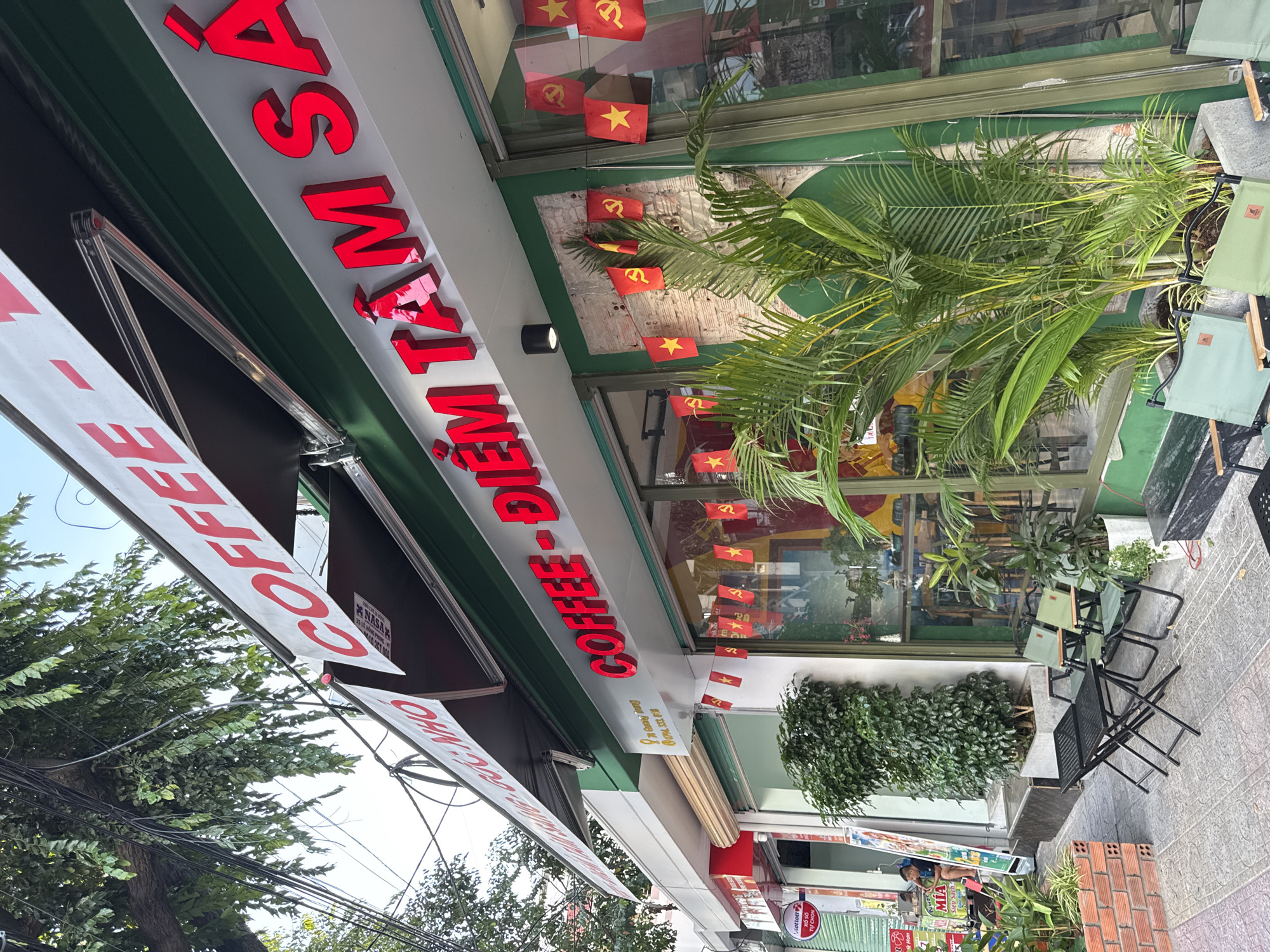 The image shows a coffee shop in Nha Trang, Vietnam. The shop's name, "COFFEE - ĐIỂM TÂM SẢ," is displayed in large red illuminated letters on the facade. The shop has a green awning and is decorated with small Vietnamese flags and banners. In front of the shop, there are several green chairs and black tables set up on a sidewalk. To the left, there is a man sitting on a stool near a tall green bush. The street is paved and lined with trees and other buildings. The sky is clear and blue.