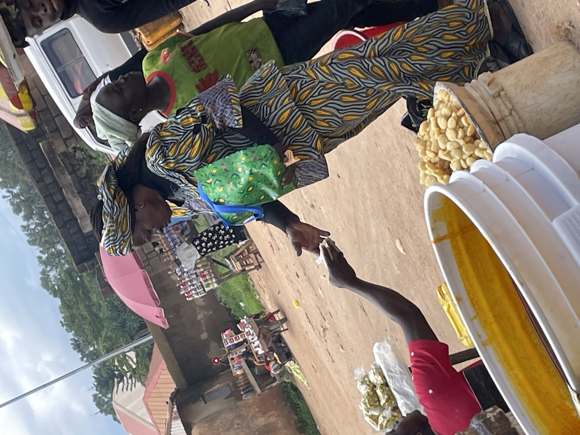 This image shows a market scene in Wang, Nigeria. The primary subjects are several individuals engaged in commerce. A person in the foreground, wearing a red shirt, is reaching out to receive something from another person. This interaction suggests a transaction is taking place.
Several objects are visible, including a large white container with yellow contents, possibly food items like groundnuts or beans, spilling out. Another white container, possibly a bucket or bowl, contains a yellow substance, likely a spice or condiment such as turmeric. There are also several smaller items packaged in plastic, and a pink umbrella is partially visible. A white truck is in the background. The ground is dirt, indicative of a market area.
The scene appears to be outdoors during the daytime, with natural light illuminating the subjects and their surroundings. The sky is visible, suggesting it is either cloudy or overcast. The setting is clearly a bustling marketplace, with various goods and people present.
The overall mood is one of activity and transaction, typical of a market environment. There is no visible text in the image.