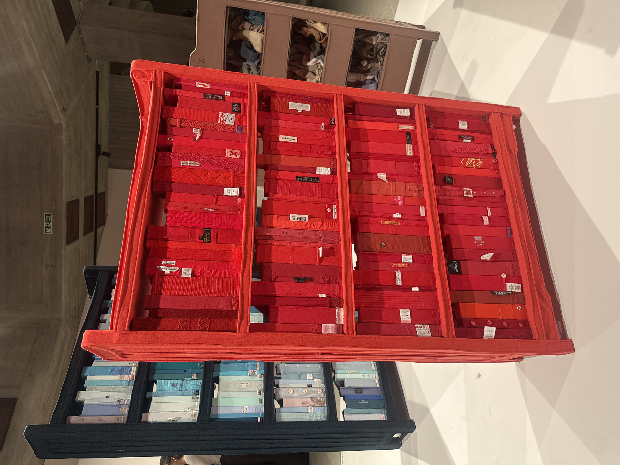 The image shows an art installation in what appears to be a gallery space. The installation consists of two fabricated structures, one stacked on top of the other. The upper structure is a bright orange or red color and is divided into compartments, each containing folded red clothing, likely shirts. The clothing in each compartment seems to be carefully arranged and stacked neatly.
Below, there's another structure, dark blue or black, also divided into compartments. These compartments are filled with folded, light blue and white clothing. The background shows an architectural space with what appear to be concrete walls and other elements typical of a modern gallery.