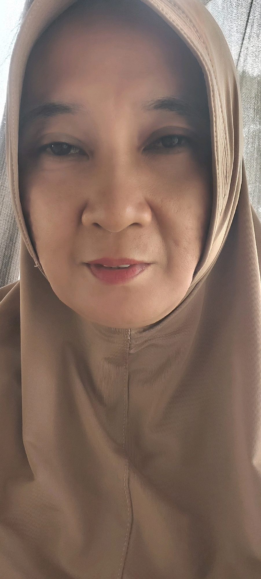The image is a close-up portrait of a middle-aged woman wearing a light brown hijab. The woman's face is illuminated by natural light, and she is looking directly at the camera with a neutral expression. Her skin appears smooth, with subtle lines around her eyes and mouth. She has dark, almond-shaped eyes and her eyebrows are well-defined. Her lips are slightly parted, revealing a hint of pink lipstick. The hijab is made of a soft, flowing fabric that drapes around her head and neck. The background is blurred, suggesting an outdoor setting, possibly a balcony or a garden, with a textured surface, possibly a wall or a curtain, visible to the right. The image captures a moment of quiet contemplation or perhaps a casual selfie, typical of personal photography. The woman's attire and appearance are consistent with Muslim women in Indonesia, and given the request for a specific location, this image could have been taken in Tanjung Redeb, Indonesia, a city known for its predominantly Muslim population and cultural practices.