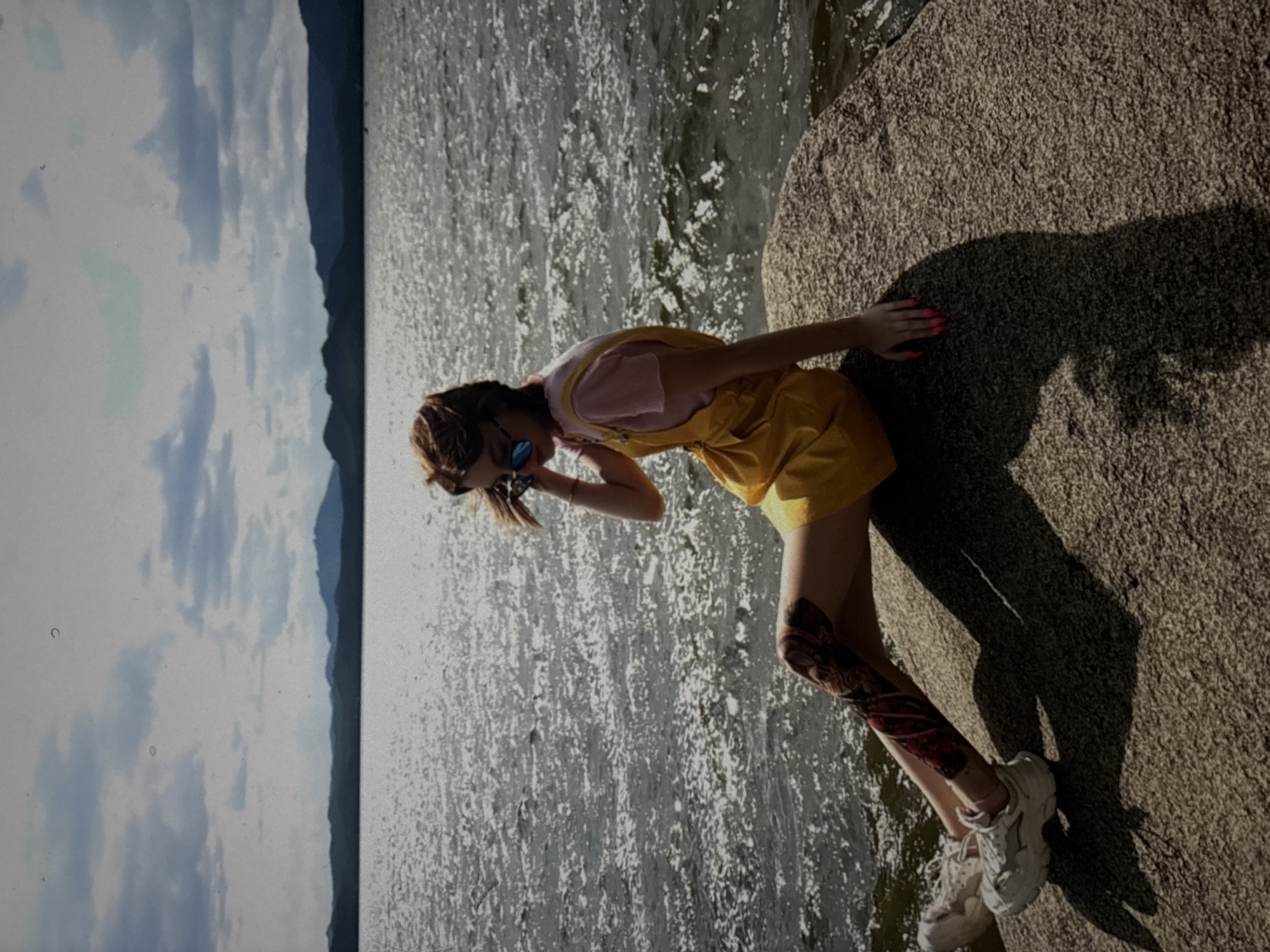 A young woman with brown hair styled in a braid is posing on a large, textured rock next to a body of water. She is wearing a pink t-shirt under a bright yellow overall dress. The dress has a bib front with straps that go over her shoulders. She has on white athletic shoes and her legs appear to have tattoos or some form of body art on them. The woman is looking down and to her left, with one hand near her face and the other extended towards the rock. Her fingernails are painted bright pink.
The water behind her is choppy and reflects the sunlight, creating a sparkling effect. In the background, there is a hazy silhouette of mountains or hills against a sky filled with white and blue clouds. The lighting suggests it is daytime, likely sunny. The overall scene appears to be a natural outdoor setting, possibly a lake or large river. The woman's pose and the bright clothing give a relaxed and casual feel to the image. No text is visible in the image. The location context is Omsk, Russia.