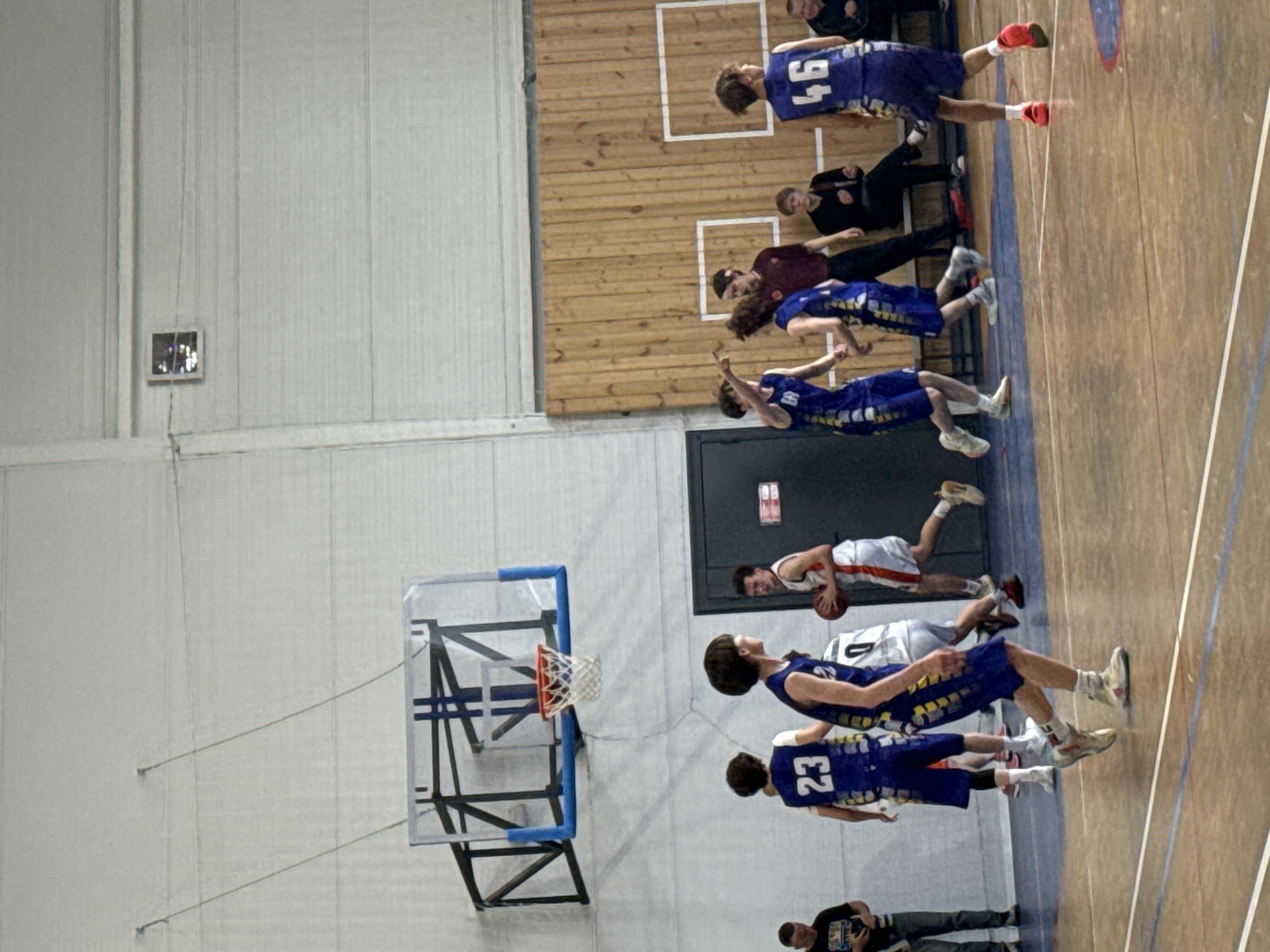 The image shows a basketball game being played in an indoor gymnasium. The players are wearing basketball jerseys with numbers on them. A basketball hoop is visible on the left side of the frame. Spectators are sitting along one side of the court. The gymnasium has a wooden floor and white walls. A door with a red sign is on one side of the court. A person is taking a photo with their phone in the lower left corner. The location is the city of Votkinsk, Russia.