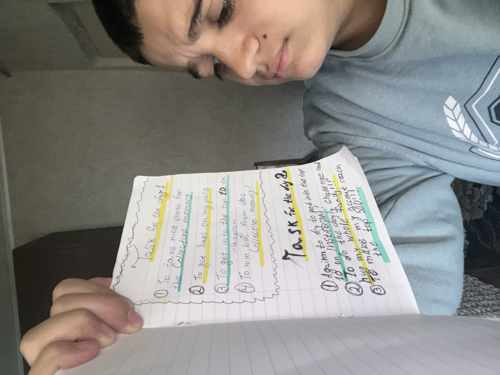 The image shows a person holding a notebook with two handwritten lists titled "Task for the day 1" and "Task for the day 2". The tasks listed seem to revolve around social media engagement, specifically on Instagram, as well as personal goals related to family interaction. The person is partially visible, focused on the notebook. The background is a dimly lit interior with a wooden cabinet visible on the left. I do not have enough information to determine if this photo was taken in Lozarevo, Bulgaria.
