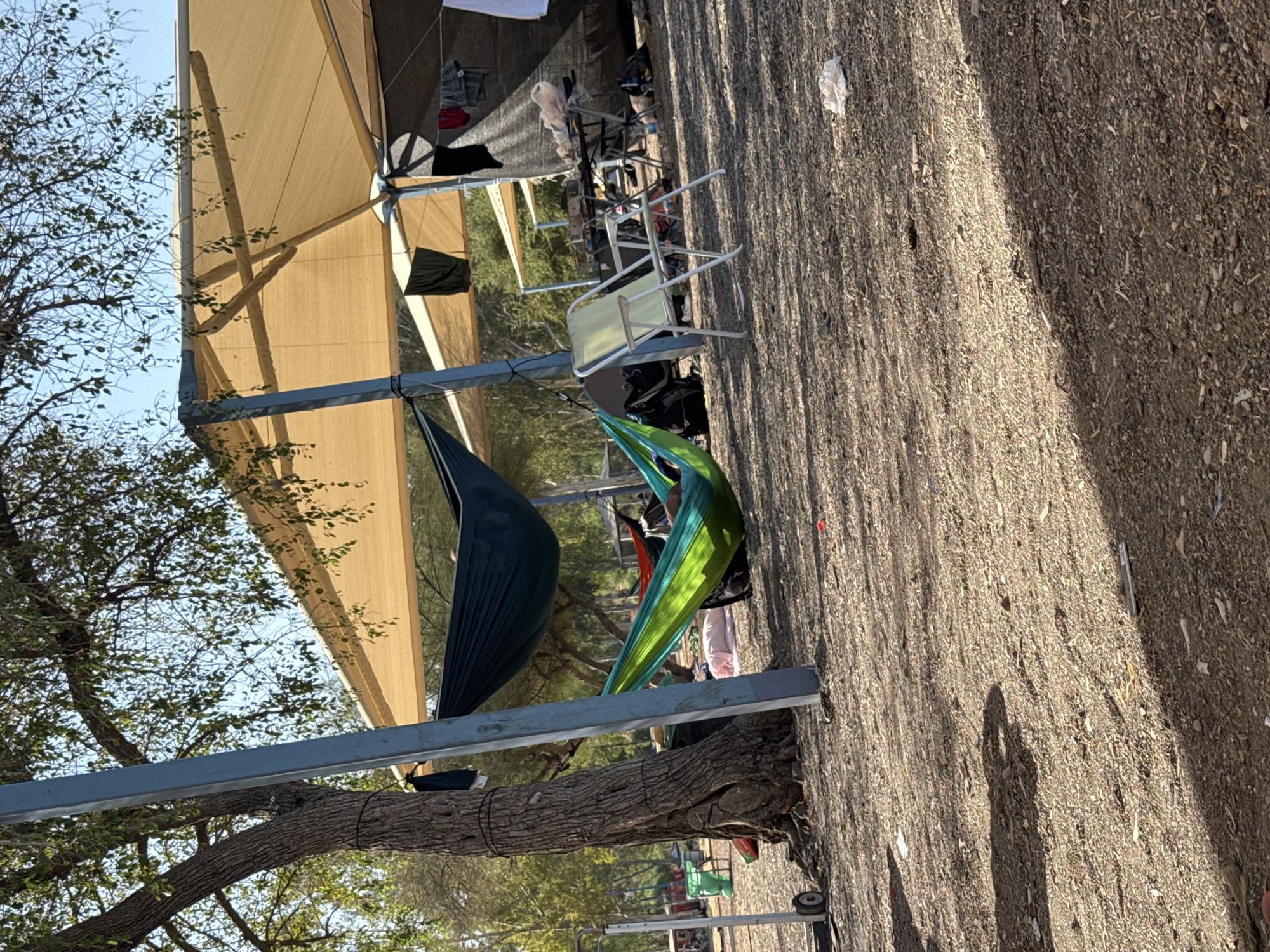 In the city of Golan Regional Council, Israel, a campsite is set up outdoors, featuring a large tree with a blue support pole attached to its trunk. Two hammocks are visible: a dark green one is suspended from the tree and the pole, and a green and blue striped hammock is also hung, with a person partially visible lying in it. Several folding chairs are present, including a light yellow one in the foreground and others in the background. A table with items on it, possibly food or cooking supplies, is set up under a tan-colored awning supported by poles. Clothing items are hung on a clothesline extending from the awning structure. The ground is covered in gravel and scattered debris, and shadows from trees and structures create a dappled pattern across the scene. The overall environment suggests a temporary outdoor living or resting area.