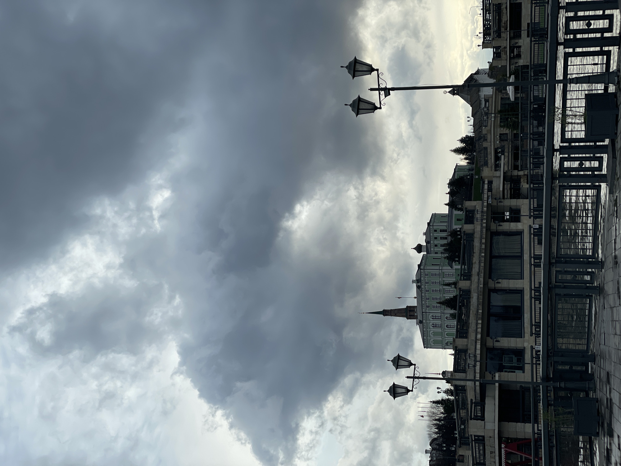 The image displays an urban landscape under a heavily overcast sky, likely viewed from a street or embankment. The photo is rotated ninety degrees clockwise; when viewed upright, the horizon runs horizontally across the lower portion of the frame, with the sky dominating the upper two-thirds.
The sky is filled with dramatic, thick clouds ranging from dark, ominous grey to brighter, diffuse white patches where the sun attempts to break through. The uneven lighting suggests dynamic weather conditions.
In the lower portion of the frame, a row of buildings stretches horizontally. A prominent light green building with white architectural details, featuring a tall, pointed spire and a rounded dome, stands out in the mid-ground. This building is characteristic of structures found within the Kazan Kremlin, specifically resembling the Governor's Palace and Söyembikä Tower, aligning with the provided location context of Kazan, Russia. Further foregrounded, there's another multi-story building with dark-framed windows and what appears to be a balcony or outdoor area.
Several ornate, dark metal streetlights or lampposts with double lanterns are visible, extending upwards from the right side of the foreground and mid-ground. Dark metal railings or fencing run along the immediate foreground, bordering a paved surface that appears wet or reflective, suggesting recent rainfall. Dark green trees are visible behind some of the buildings.
The time of day appears to be daytime, possibly late morning or afternoon, given the variation in cloud cover and light. The overall mood conveyed by the heavy clouds is somewhat dramatic or somber. There is no visible text in the image.