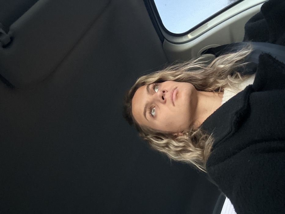 A young woman with wavy blonde hair and green eyes looks up, seemingly from inside a car. She is wearing a black coat over a light-colored top. The car's interior is visible, including the sunroof and the sun visor. The angle of the shot is from below, emphasizing the woman's upward gaze. The background is mostly dark.