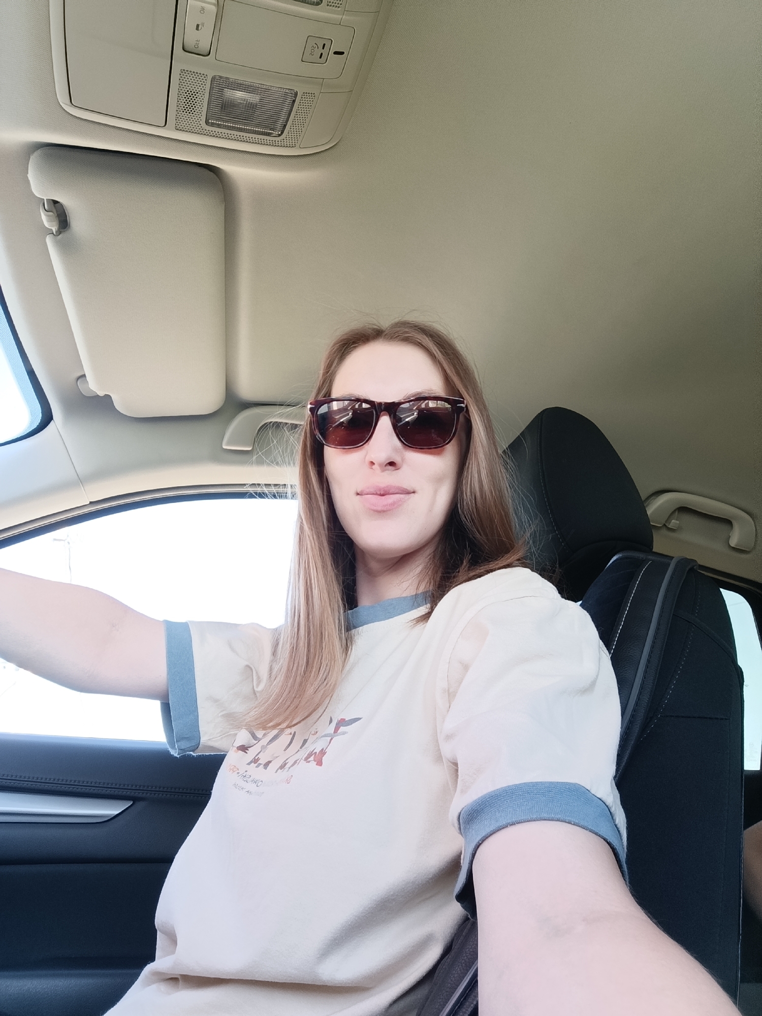 A woman is sitting in the driver's seat of a car. She is wearing sunglasses and a t-shirt. The car's interior is visible, including the dashboard, steering wheel, and seats. The location appears to be outside, based on the view through the car window. There is no information in the image to confirm the city of Chaikovsky, Russia.