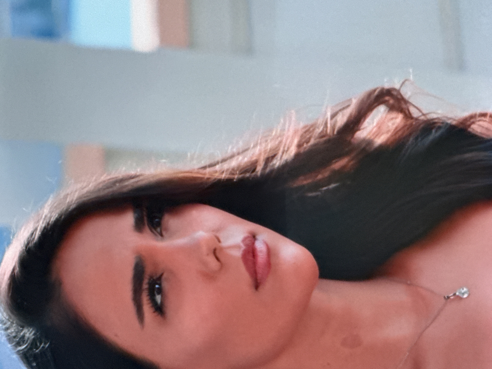 A close-up, head-and-shoulders shot shows a young woman with long, dark brown hair parted to her left. Her skin has a warm, reddish tone. She has prominent eyebrows, dark eyeliner, and her eyes are looking to her left. Her lips are full and painted with a pinkish-red lipstick. A light-colored pendant necklace with a single tear-drop shaped gem is visible on her neck, on the left side of her collarbone. The background is out of focus and appears to be a light blue sky with hints of white and pink structures, suggesting an outdoor setting during the day. The woman is likely in the city of Uyutnoye, Ukraine.