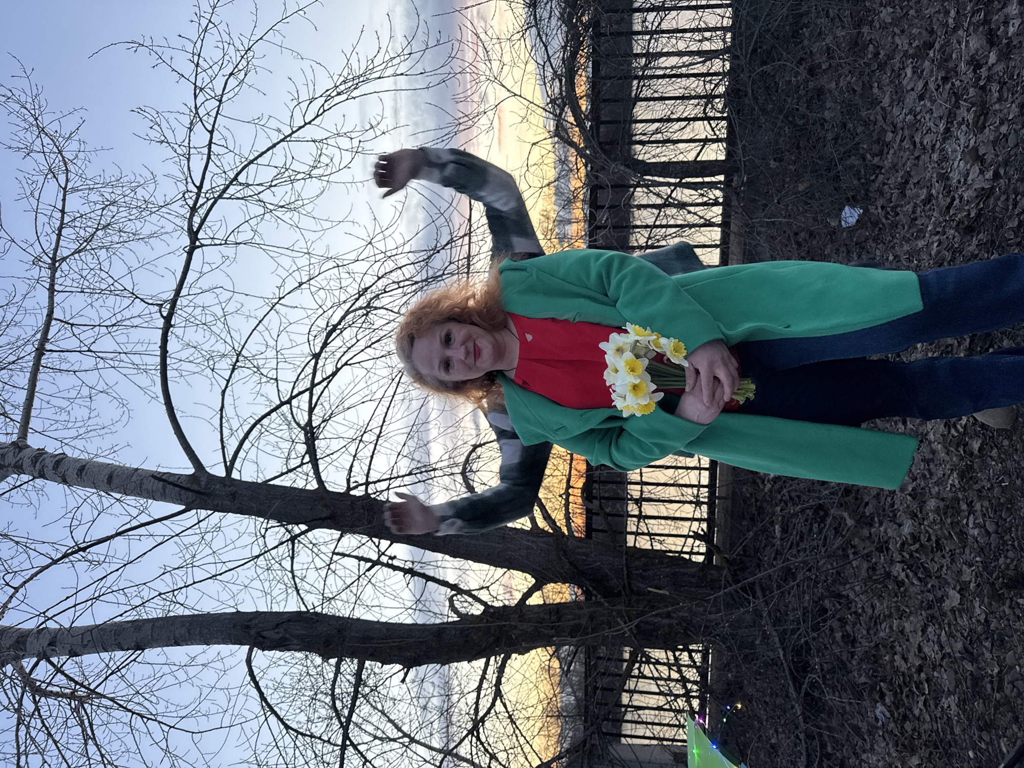 A woman with reddish-blonde hair and a light complexion stands outdoors, smiling gently at the camera. She is wearing a bright green coat over a red top and dark blue jeans. In her hands, she holds a small bouquet of white and yellow daffodils. Partially visible behind her is another individual, wearing a dark plaid or striped long-sleeved shirt, with one arm raised in the air.
The setting is an outdoor area, likely a park or natural space, characterized by numerous tall, bare trees with slender branches, indicating late winter or early spring. A dark, slatted fence or railing runs across the middle ground, separating the foreground from the background horizon. The sky transitions from a light blue overhead to warm hues of yellow, orange, and soft pinks near the horizon, suggesting sunset or dusk. The ground beneath is covered with fallen, dark leaves and natural debris. The atmosphere appears calm and pleasant. There is no visible text in the image. The location context is Chaikovsky, Russia.