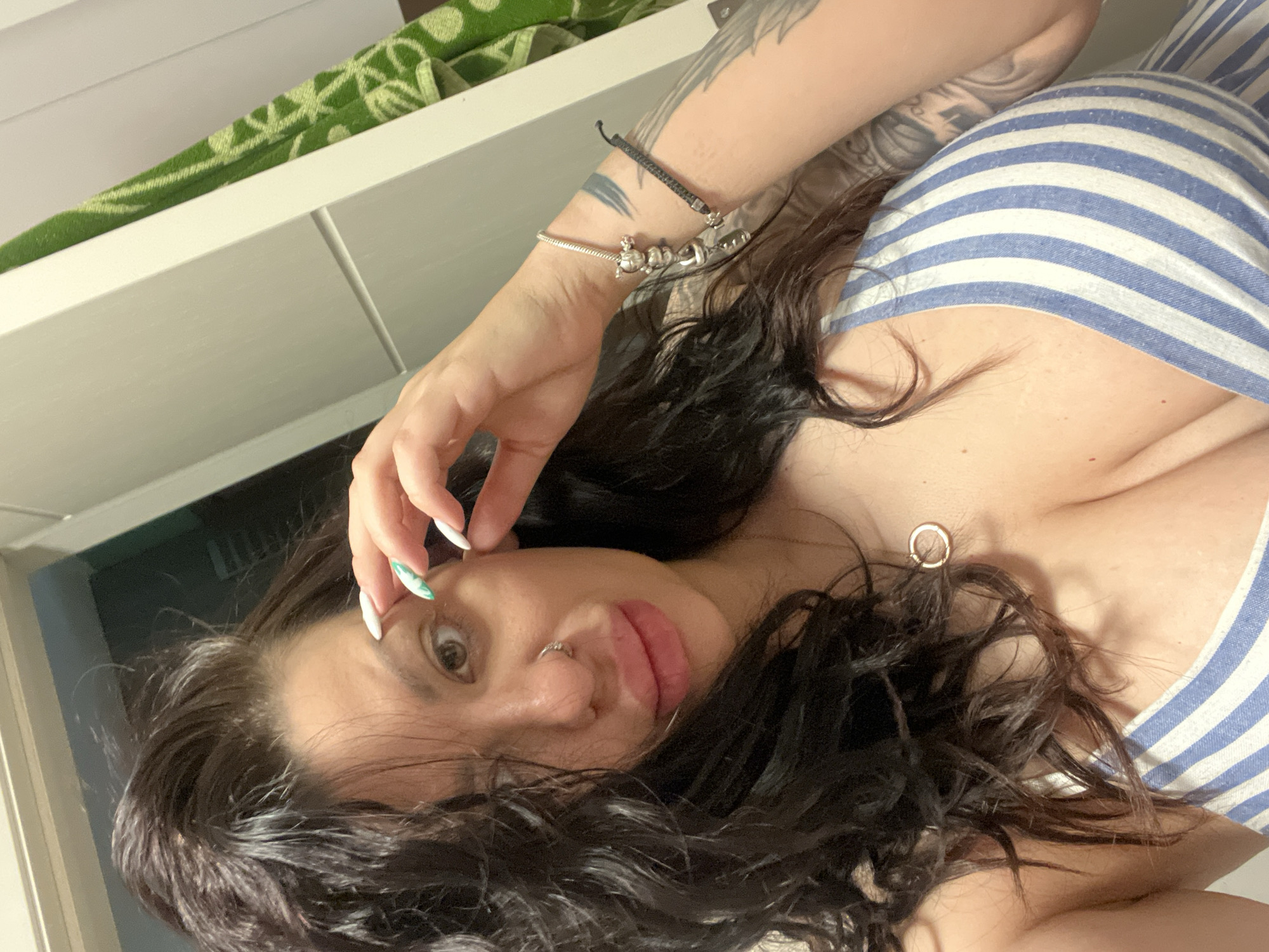 A woman with long, dark, wavy hair is lying down, facing the camera. She is wearing a blue and white striped top. Her left hand is raised near her face, showcasing white and green nail polish. A silver bracelet and a cord bracelet are visible on her left wrist. A small nose ring is in her left nostril. A circular pendant hangs from a delicate chain around her neck. The background includes a white surface, possibly a shelf or cabinet, and a green patterned object. The image does not provide any information about the location of Plovdiv, Bulgaria.