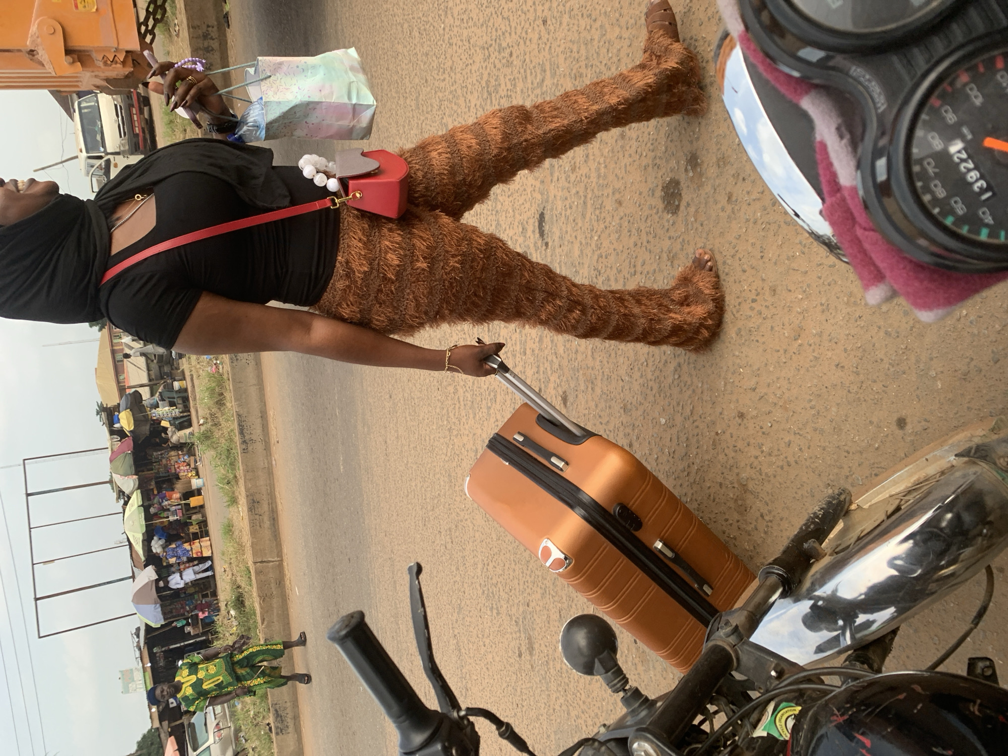 A person walks on a paved road in Shagamu, Nigeria, pulling an orange suitcase. The person is wearing a black headscarf, a black short-sleeved shirt, and shaggy brown pants. They are also carrying a red handbag over their shoulder and holding a shopping bag. A motorcycle is partially visible in the foreground. In the background, there are market stalls with umbrellas and people shopping. A white van and a large orange vehicle are also present.