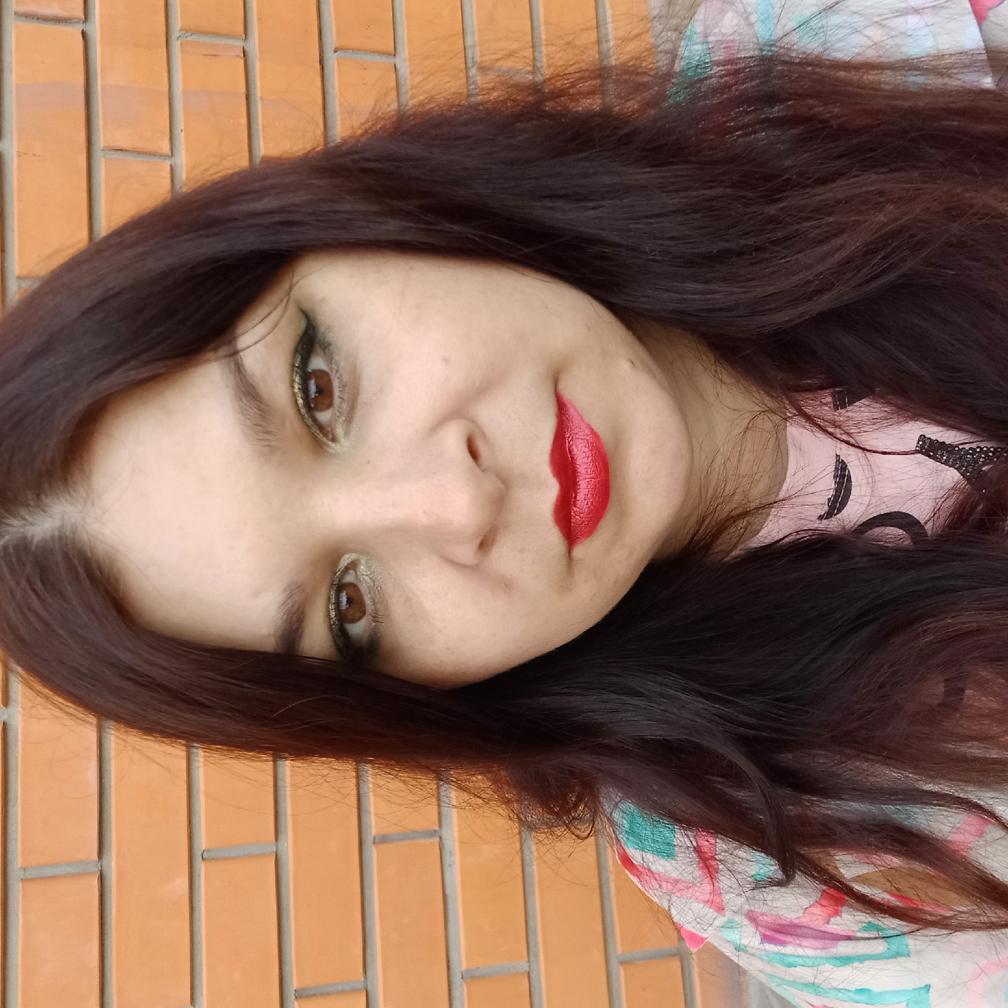 The image is a close-up selfie of a woman with dark brown hair and brown eyes, taken outdoors in front of a brick wall. She is wearing makeup, including eyeshadow and bright red lipstick. Her clothing, a pink t-shirt with a design and a colorful patterned shirt over it, is partially visible. The brick wall appears to be an exterior wall of a building. There are no other people or objects of significance in the frame. The lighting suggests it is daytime. The scene is static and there is no visible action. No specific landmarks or identifying features of Kropyvnytskyi, Ukraine are discernible in this image.