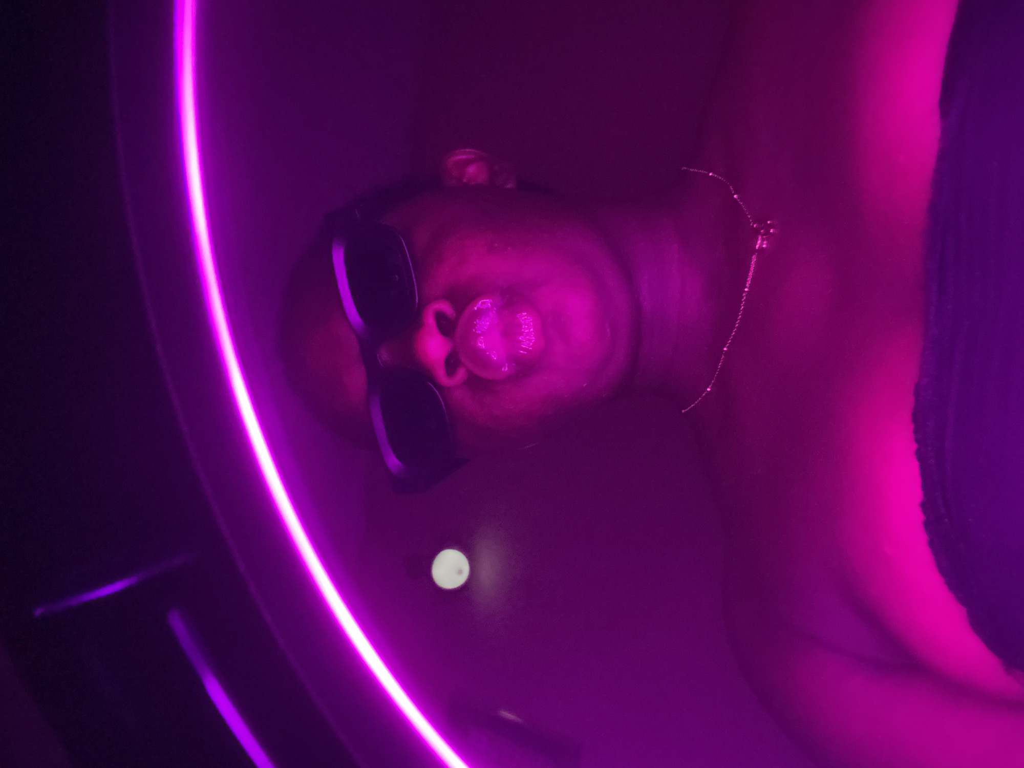 A close-up, canted, aerial shot features a person immersed in a round tub or vessel illuminated with a strong magenta/purple light. The person, wearing dark sunglasses and a thin necklace, is seen from the nose upwards, blowing a bubble. The light is intense, giving the skin a magenta cast. Part of the person's body and what appears to be a black garment can be seen to the right of the frame. A bright, circular reflection is visible in the water. It is not possible to determine the location in Maigizo, Nigeria or any specific actions or interactions due to the perspective and limited context.