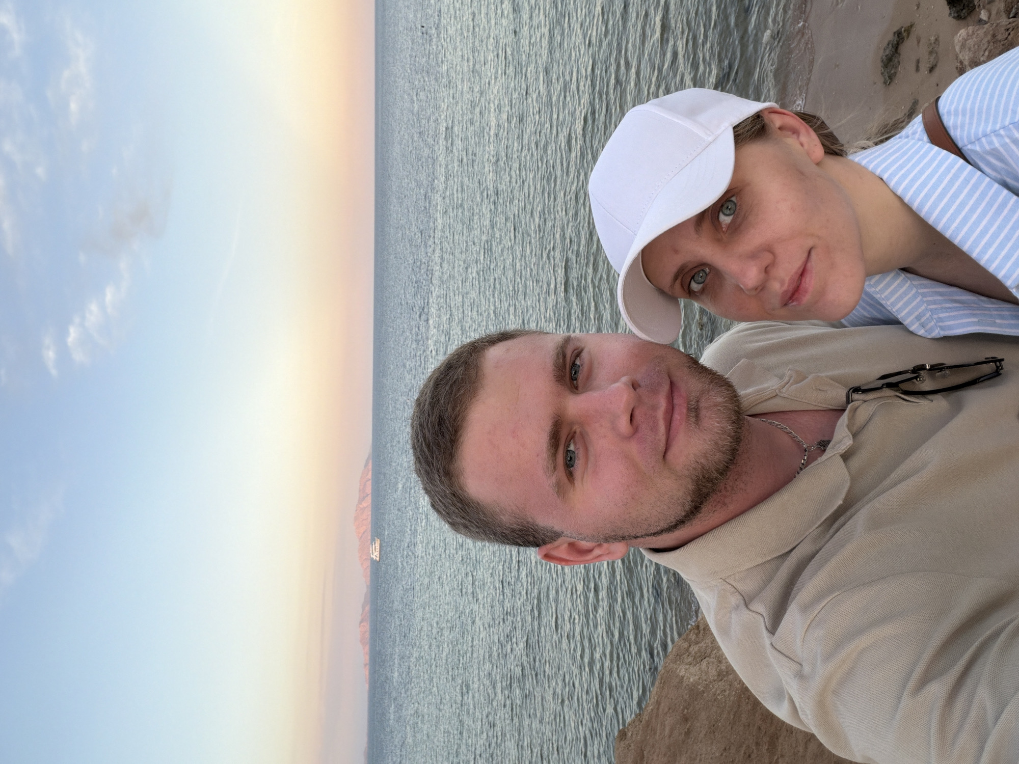 An outdoor portrait depicts two individuals positioned on a sandy and rocky incline adjacent to the Red Sea, consistent with the coastline of Sharm El Sheikh, Egypt. The scene is illuminated by the soft light of twilight, exhibiting a sky gradient from light blue to soft orange and pink near the horizon.
In the foreground, a male with short, light brown hair, light stubble, and blue eyes is situated to the left, wearing a light beige polo shirt with sunglasses hanging from the collar and a silver chain visible around his neck. To his right and slightly behind him is a female with light-colored hair, blue-green eyes, wearing a white baseball cap and a blue and white striped button-up shirt; a brown strap is visible over her right shoulder. Both individuals are looking directly at the camera with neutral expressions.
The midground consists of the calm, rippled surface of the Red Sea. A white motorboat is visible on the water, positioned equidistant between the foreground coastline and the distant horizon.
The background features several prominent, reddish-brown landmasses or islands, consistent with Tiran or Sanafir islands, silhouetted against the horizon. The sky overhead is light blue with sparse, wispy clouds.