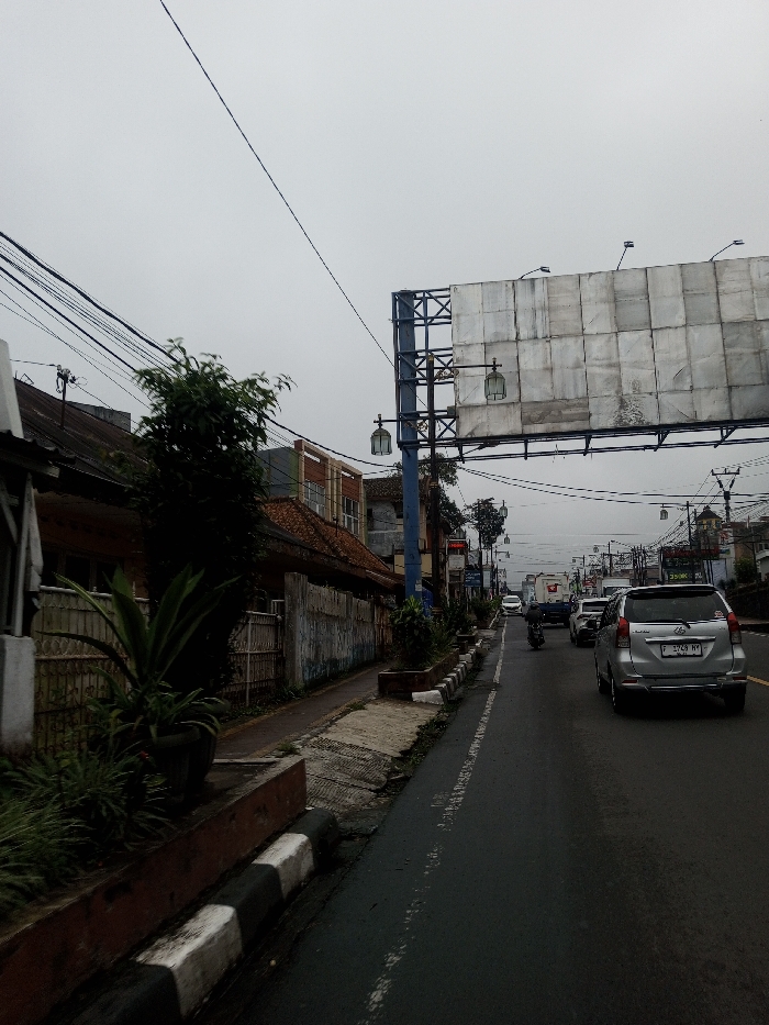 The image displays an overcast daytime street scene. A multi-lane asphalt road occupies the center and right, featuring a faded white dashed line and black-and-white striped curbs. A silver Toyota Avanza (license plate F 1749 NY) is positioned on the right lane, facing away from the viewer, with other vehicles and a motorcycle visible in the traffic stream further down the road.
To the left, a concrete sidewalk runs parallel to the road, bordered by a dark brown planter box containing green foliage, including potted plants. Beyond the sidewalk, a metal fence precedes a series of older, low-rise buildings with red tiled roofs and varied facades, obscured by dense utility wiring in the upper left.
On the right side of the road, a large, unadorned metal billboard structure is mounted on a blue support pole. The billboard is equipped with multiple upward-facing floodlights. Below it, two decorative streetlights with rectangular lanterns are affixed to the same pole. Additional utility poles with numerous power lines and distant buildings line both sides of the street under a uniformly gray sky. No discernible individuals are present.