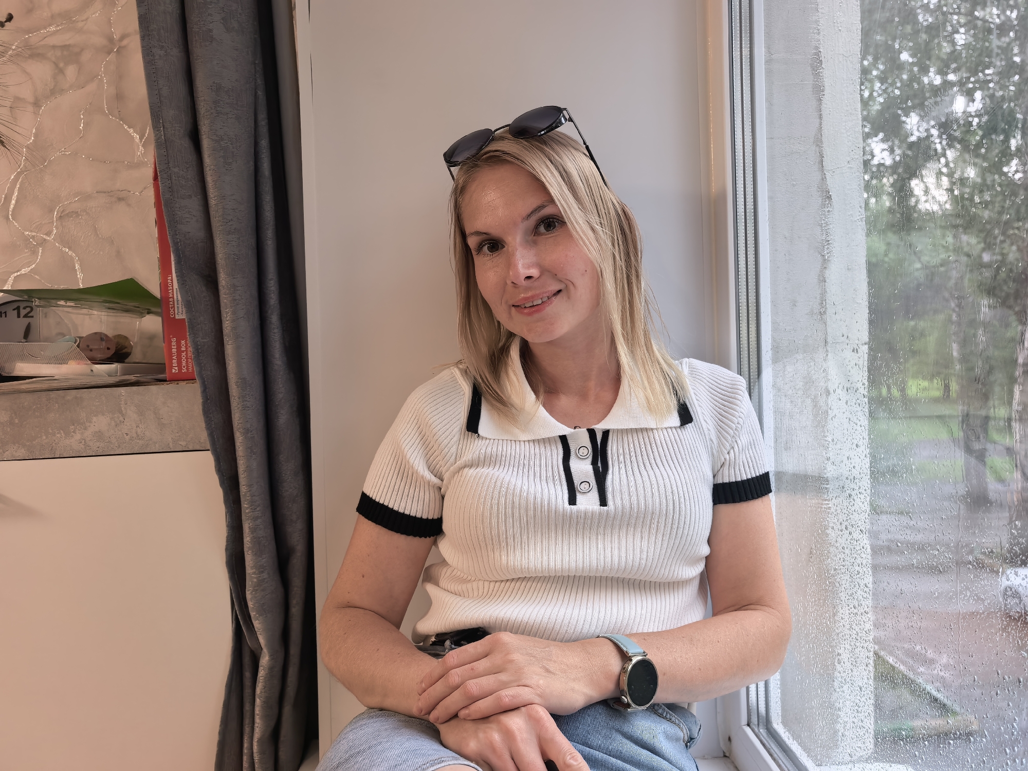 A blonde woman with sunglasses perched on her head sits by a window. She is wearing a white short-sleeved collared shirt with black trim and blue jeans. Her hands are clasped in her lap, and she is wearing a light blue smartwatch on her left wrist. Outside the window, it appears to be raining, with water droplets visible on the glass and trees in the background. To the left of the woman, a gray curtain hangs, and a shelf with various items, including what appears to be a red box and a clear container with small objects, is visible. The overall scene suggests a domestic setting. This image is not located in Sterlitamak, Russia.