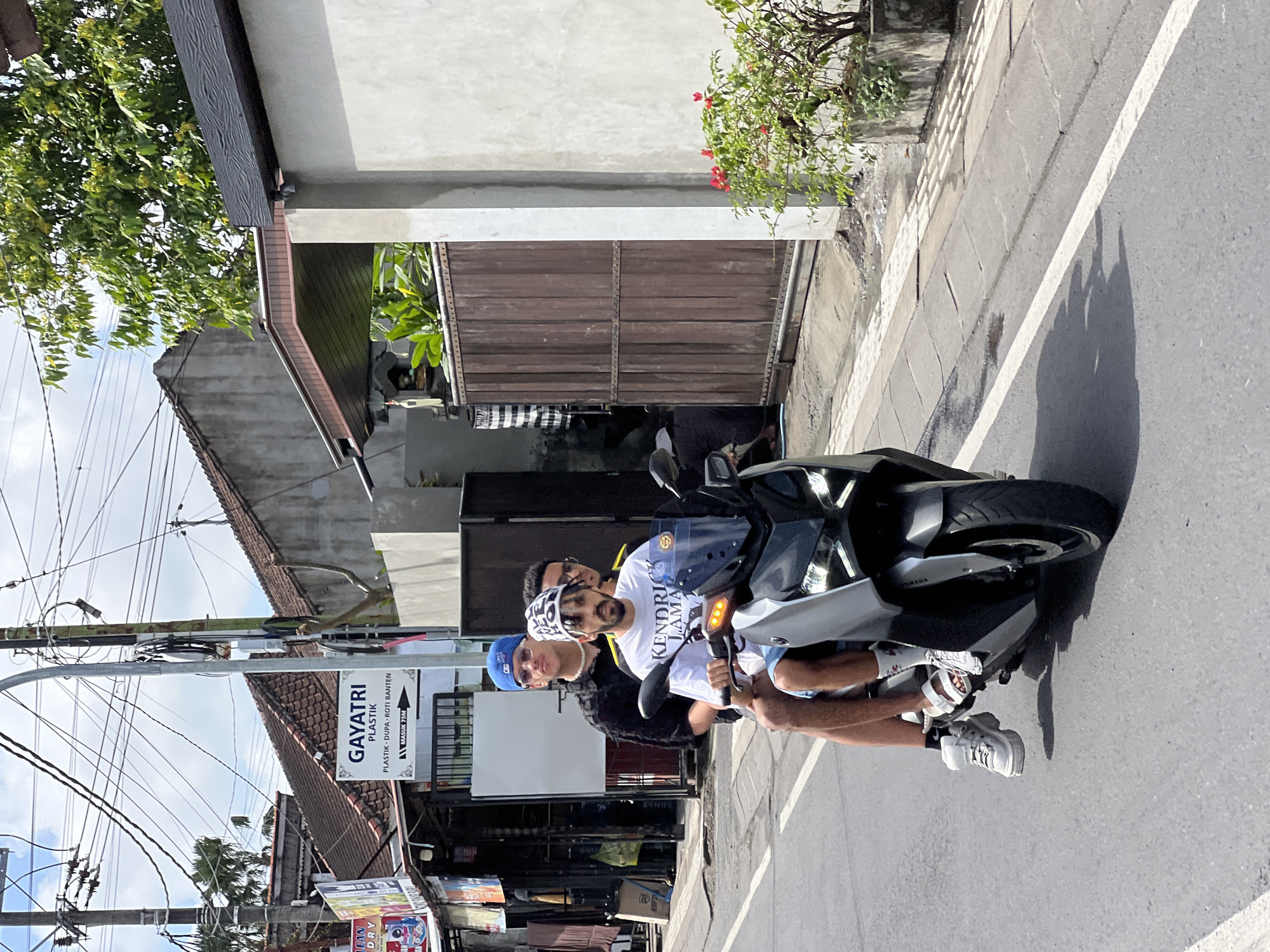 Three individuals are riding on a gray scooter on a paved road in North Kuta, Indonesia. The person in the front is wearing a white t-shirt with "Kendrick Lamar" printed on it, a white hat with black lettering, and white sandals. The person in the middle is wearing a black shirt, sunglasses, and a blue baseball cap. The person in the back is partially obscured, wearing glasses and a yellow shirt. They are riding on the right side of the road. Buildings with tiled roofs and a wooden gate are visible along the right side of the road. Electrical wires crisscross overhead. A sign with "GAYATRI PLASTIK" is visible on a building to the left. The sky is partly cloudy.