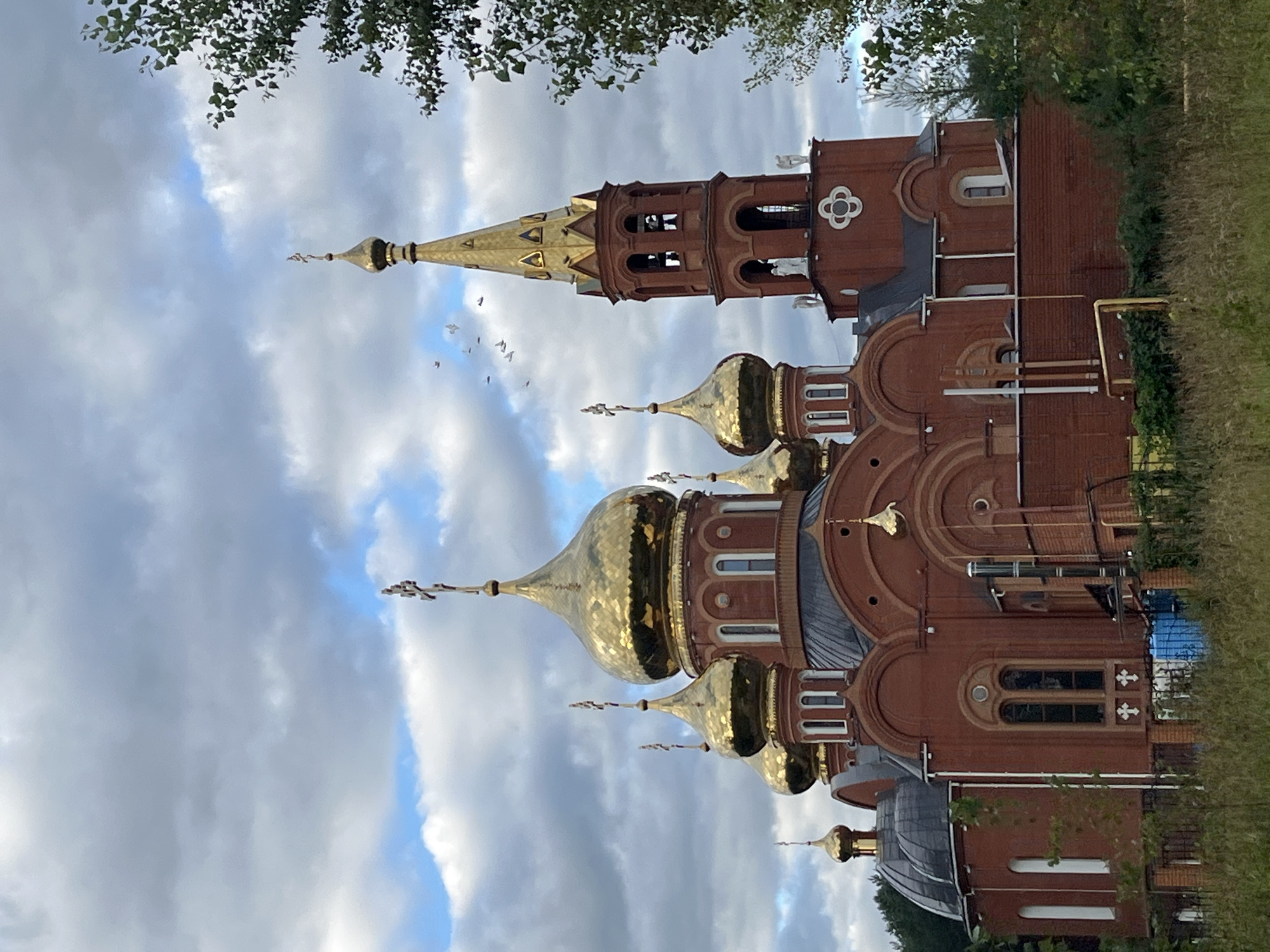 The image shows the Resurrection Cathedral, a Russian Orthodox church in the city of Chaikovsky, Russia. The cathedral is a large, brick building with multiple gold-domed cupolas and a tall, steeple-like bell tower. The architecture is typical of Russian Orthodox churches, with a red brick exterior and ornate golden domes topped with crosses. The sky is cloudy with patches of blue sky visible. Trees and green vegetation surround the church, suggesting it is located in a somewhat rural or suburban area. The foreground is filled with dry grass and some fencing. The overall impression is of a majestic religious building set against a dramatic sky.