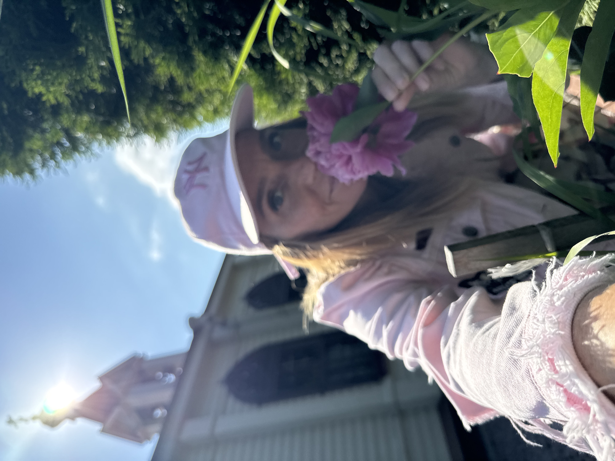 A young woman is pictured outdoors, holding a pink flower up to her face. She is wearing a pink baseball cap with an "NY" logo and a pink denim jacket with distressed edges. The background shows a bright blue sky with the sun's rays shining through, and the blurred image of a building with a steeple, likely in Lomonosov, Russia. The overall impression is of a sunny day and a casual, cheerful moment. The perspective is from below, looking up at the woman and the sky.