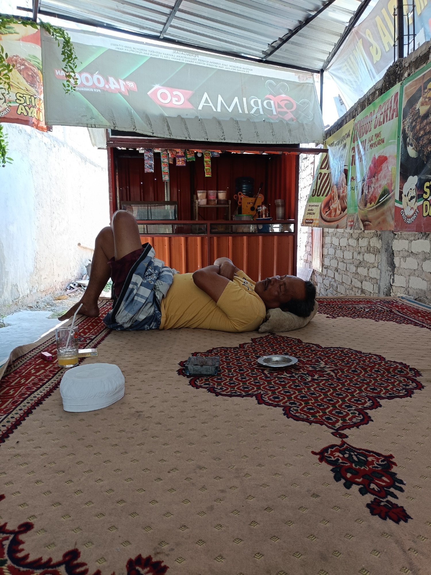 A man in a yellow shirt and plaid shorts sleeps on a large carpet. He is lying on his back with his arms crossed over his chest and his head on a pillow. A glass of orange juice and a pack of cigarettes are on the carpet near his head. A white peci, or cap, is also on the carpet nearby. The scene appears to be outdoors under a covered structure with banners advertising food and drinks hanging in the background. The man is positioned on a large, patterned carpet, which is laid out on a dirt or gravel surface. The backdrop includes a building with a red corrugated metal exterior and a white brick wall. The overall setting suggests a casual outdoor eatery or resting area in Pordapor, Indonesia.
