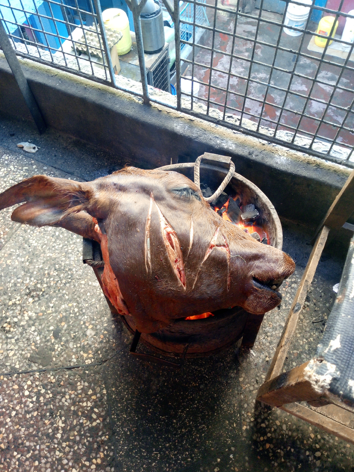 A charred animal head, likely a cow or goat, rests in a metal container filled with hot coals. The head is dark brown, with visible cuts in its skin. Flames are visible licking at the head. The container sits on a terrazzo floor, next to a metal railing and a wooden structure that appears to be a chair or table. The scene is likely taking place outdoors, possibly on a balcony or patio. The backdrop features a blue-painted metal fence and glimpses of the surroundings, which potentially indicate an urban setting. Given the prevalence of grilled meat in Kenyan cuisine, this could be a street food preparation scene in Nairobi, Kenya.
