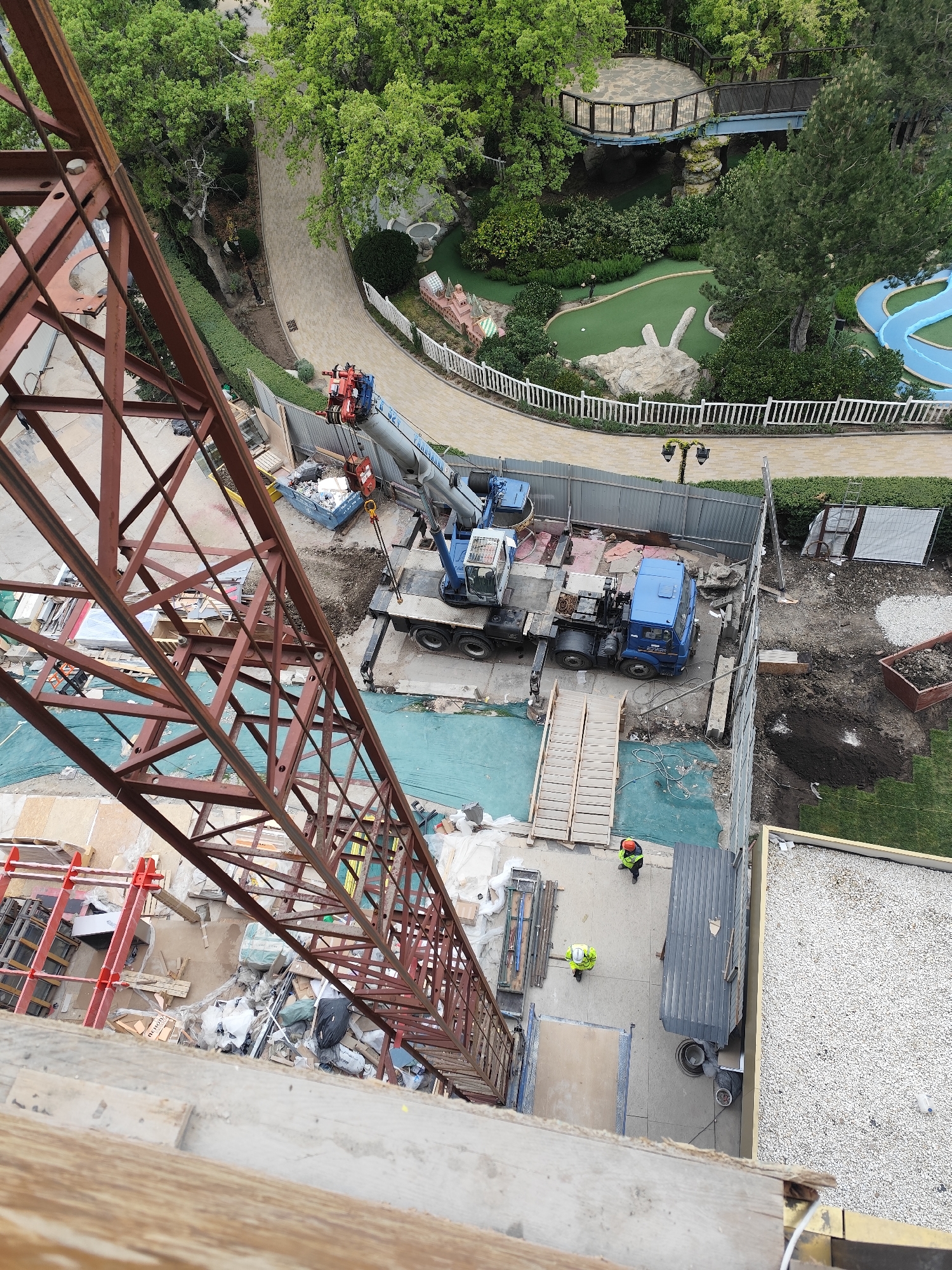 A high-angle shot presents a construction site in Симеизский поселковый совет, Ukraine. Dominating the view is a large rust-colored crane positioned centrally with its boom extending toward the top of the frame. Adjacent to the crane's base, a blue truck and a smaller crane are situated behind a temporary metal fence. Workers in neon vests are visible on the ground, and construction materials are scattered around the site.
Beyond the construction area, a miniature golf course with green turf and a winding path is seen, suggesting a recreational or resort setting. A pedestrian bridge is visible amid the lush greenery in the background.