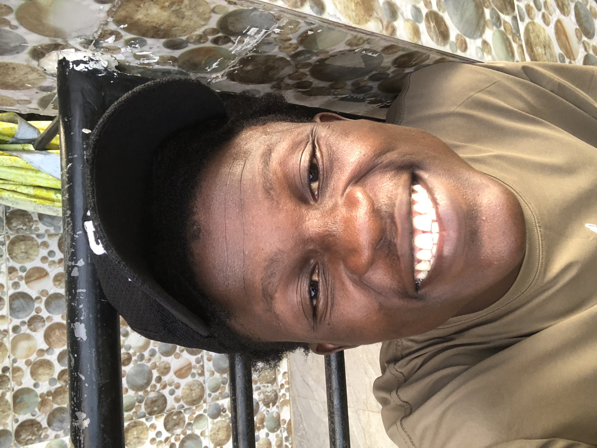 A close-up, selfie-style photograph shows a person wearing a black baseball cap and a brown t-shirt, smiling broadly at the camera. The person's face is illuminated by natural light, highlighting their features and white teeth. The background is a wall decorated with an assortment of round, mosaic-like tiles in shades of beige, brown, and gray, with a black metal railing running horizontally across the upper portion of the image. The overall impression is a casual, outdoor portrait. While the image itself does not contain specific indicators of location within Ado, Nigeria, it captures a moment of personal expression against a visually interesting backdrop.
