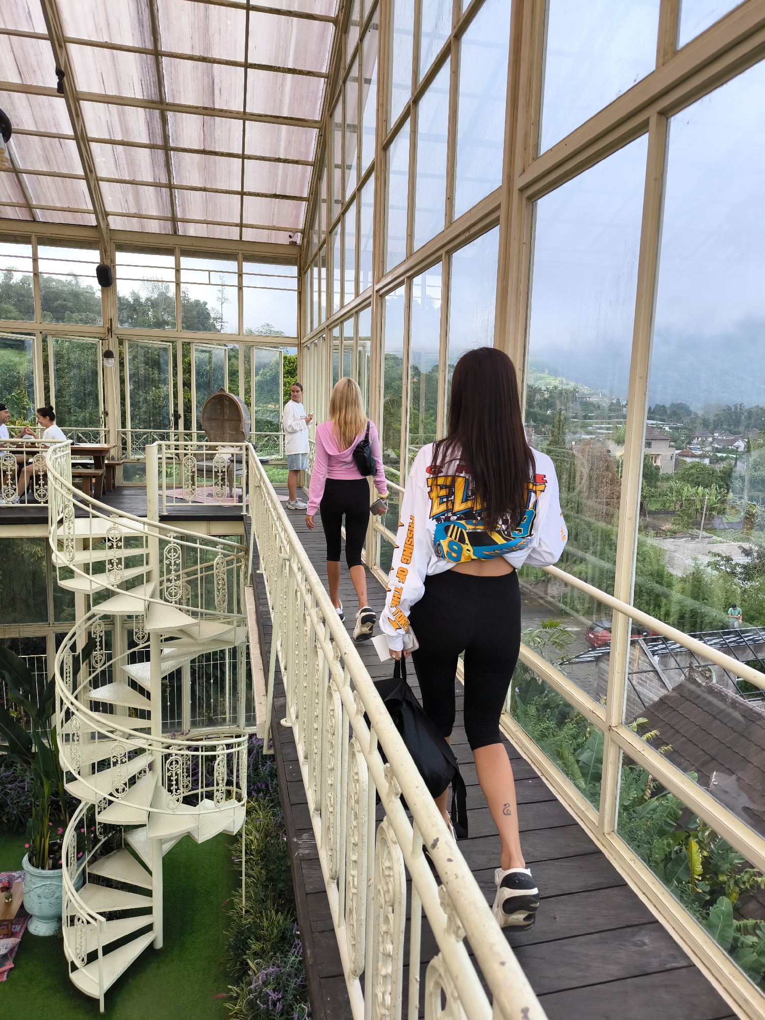 The image shows the interior of a glass-enclosed structure in Candikuning, Indonesia. Two women are walking away from the camera on a wooden bridge with a white railing, toward two other people. One woman wears a white long-sleeved shirt with a colorful graphic on the back and black leggings, carrying a black backpack. The other woman wears a pink hooded jacket and black leggings. To the left, a white spiral staircase descends to a lower level with plants. Other people are seated at a table in the background. Outside the glass walls, houses and greenery are visible.