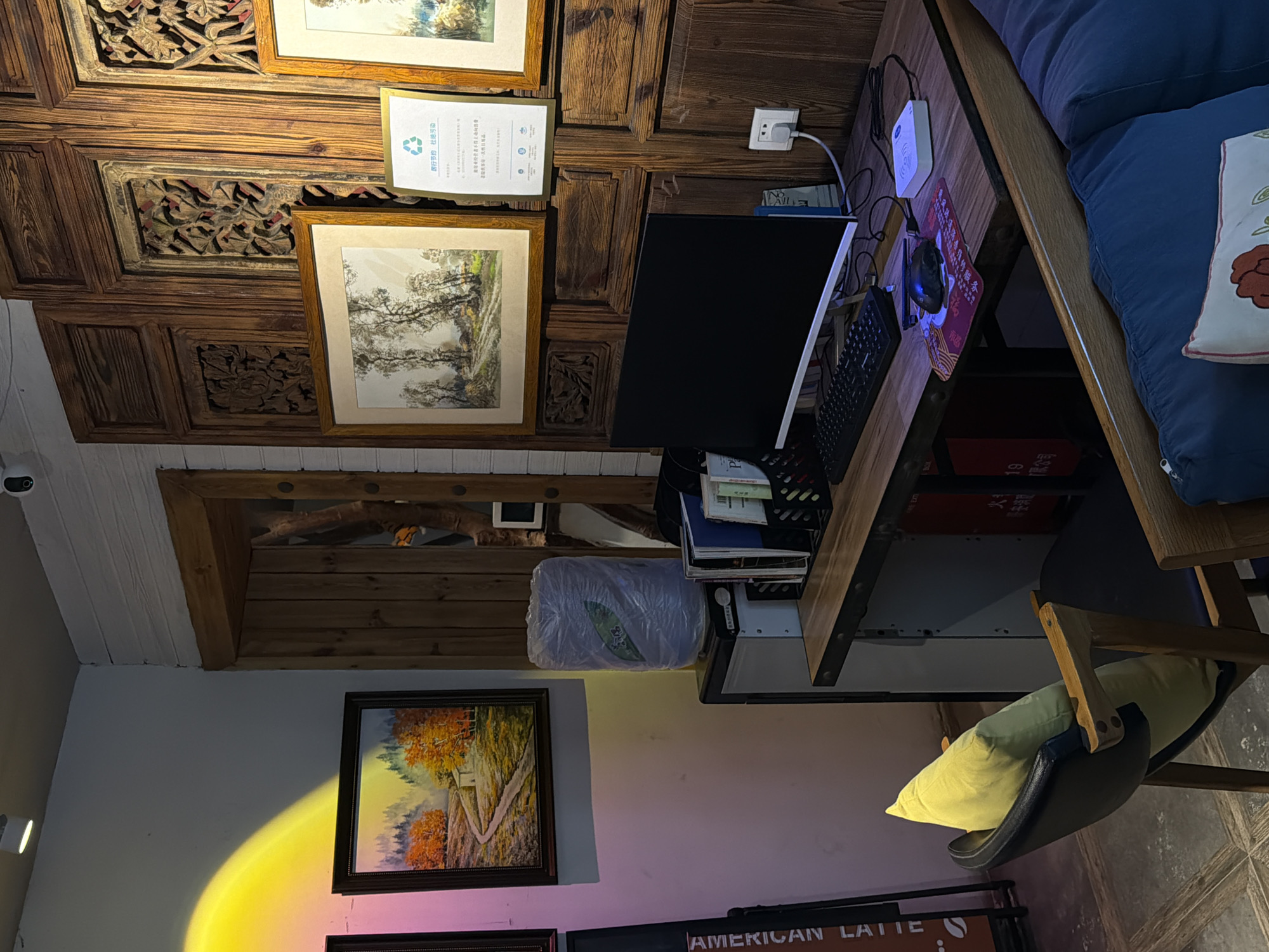 The image depicts an indoor scene, likely a café or office space, characterized by a blend of rustic and modern elements. A desk with a computer monitor, keyboard, and mouse is positioned against a backdrop of intricately carved wooden panels. To the left of the desk, a chair with a yellow pillow is visible, and beyond that, a painting of a landscape with autumn trees is illuminated by a yellow spotlight. On the desk, a wireless charging pad and a paper with recycling symbols are present. A white, cylindrical object wrapped in plastic sits on a small cabinet to the left of the desk. The environment appears to be well-lit, with a combination of natural and artificial light sources. There are no people visible in the image. The overall impression is one of a thoughtfully designed space that combines artistic elements with functional workspace.