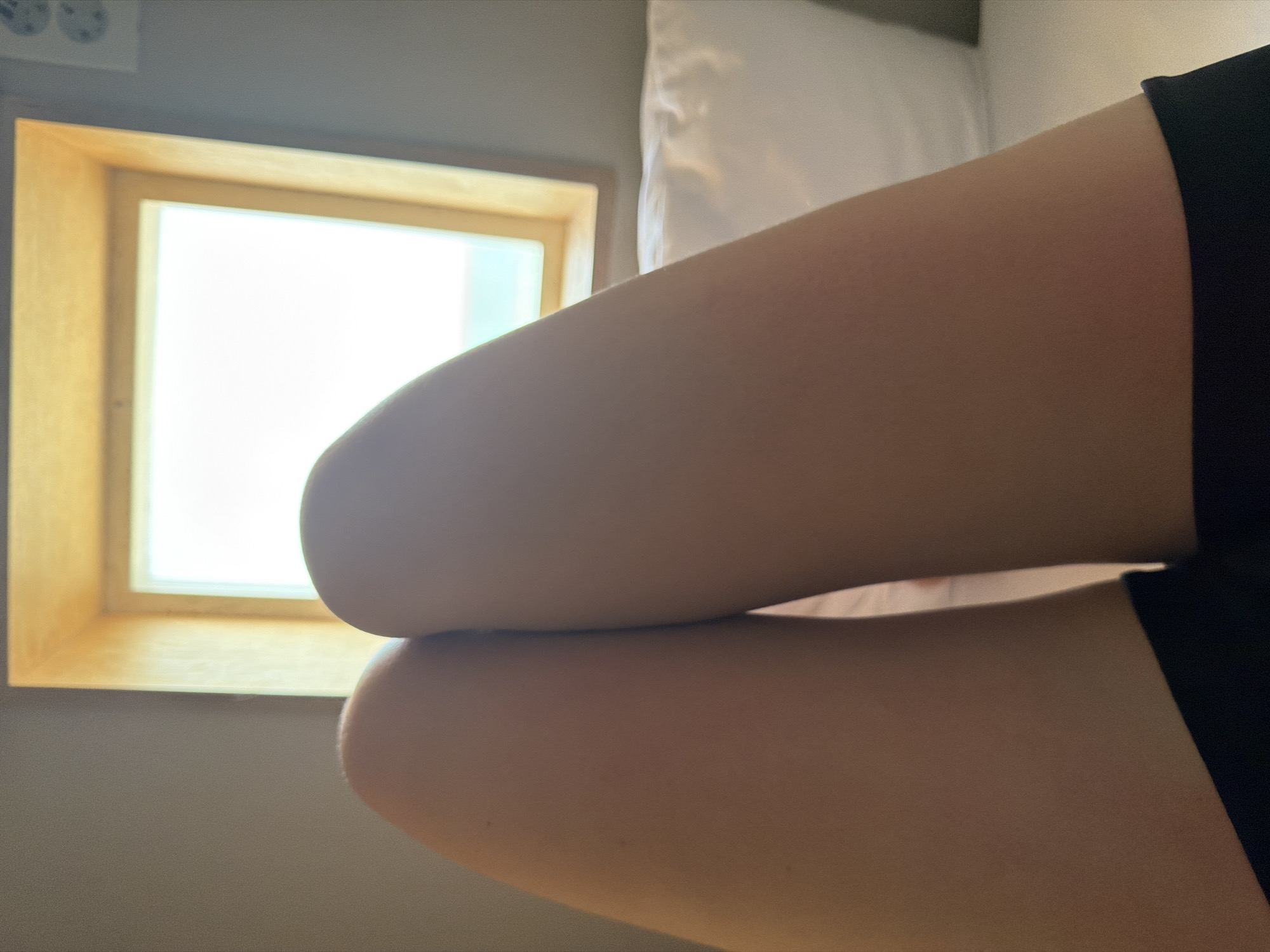 The image shows a close-up view of a person's legs, with a window in the background. The person is wearing dark clothing, possibly shorts or a skirt, at the hem of the frame. The window appears to be a skylight or a dormer window, with a wooden frame. Bright light is coming from the window, suggesting it is daytime. The surrounding wall is a neutral color. A white pillow is visible to the right of the legs. The scene appears to be indoors, likely a bedroom, with the person positioned on a bed or similar surface. There is no specific indication of the location being in Gangneung-si, South Korea.