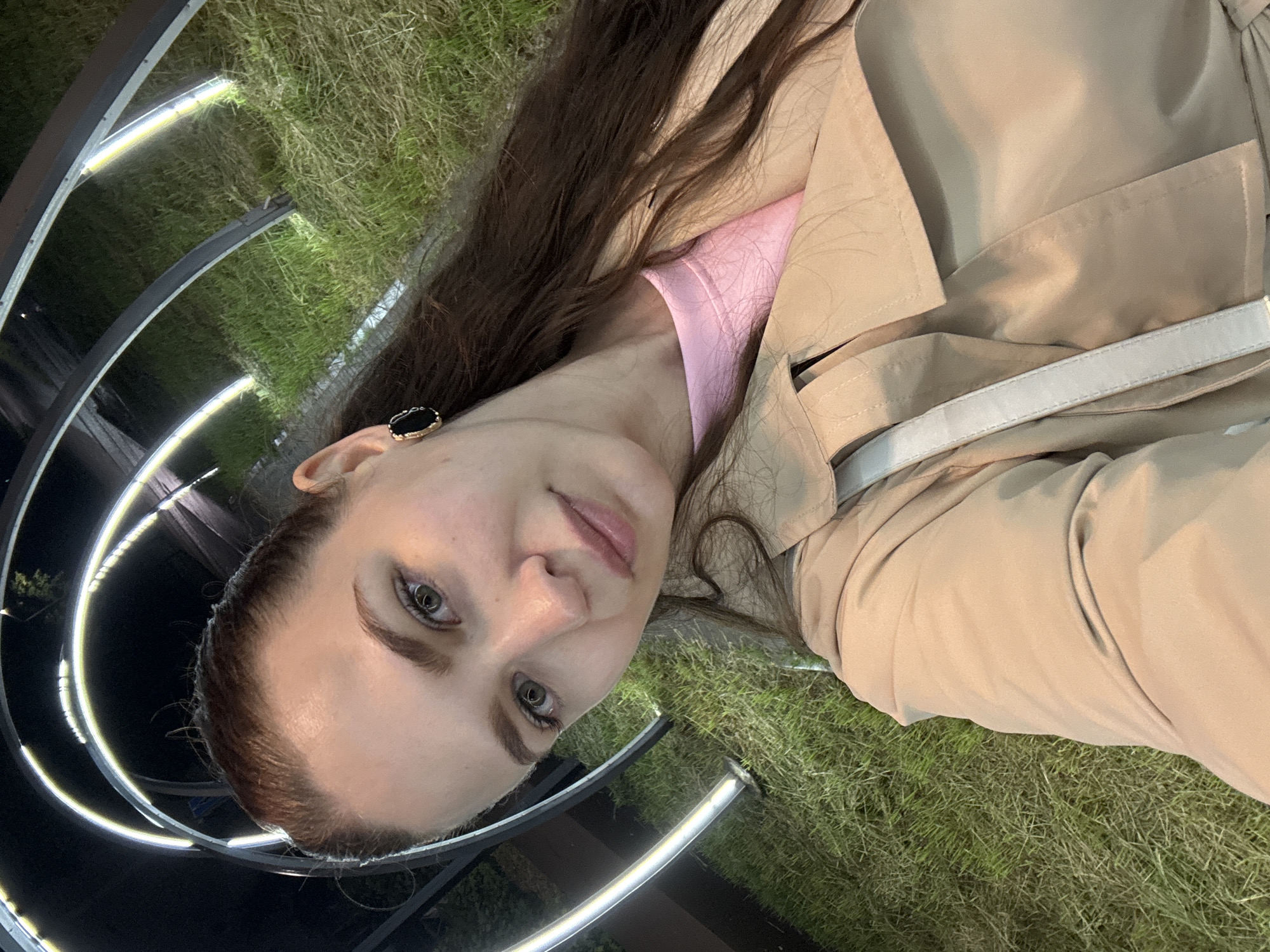 A forensic examination of the image reveals a nighttime selfie of an adult female in Chaikovsky, Russia. The subject, a Caucasian woman appearing to be in her 20s or 30s, occupies the foreground, captured from a slightly low angle. She has light skin, light-colored eyes (likely green or hazel), and long, dark brown hair styled back from her face, with strands visible over her right shoulder. She wears a light pink crew-neck garment beneath a beige or light tan trench coat. A single black earring with a gold-colored border is visible on her right ear. A light-colored strap, consistent with a shoulder bag, crosses her left shoulder and upper chest. Her expression is neutral as she looks directly at the camera.
The background is dark, indicating evening or night conditions. It features a series of interconnected, arching structures, constructed from dark material, forming an illuminated pathway. These structures are equipped with bright white LED strip lights along their inner curves, casting illumination onto the ground. The ground beneath these structures is covered in natural grass, displaying variations in texture and color, consistent with an outdoor public space or park. No other individuals or specific landmarks are discernible.
