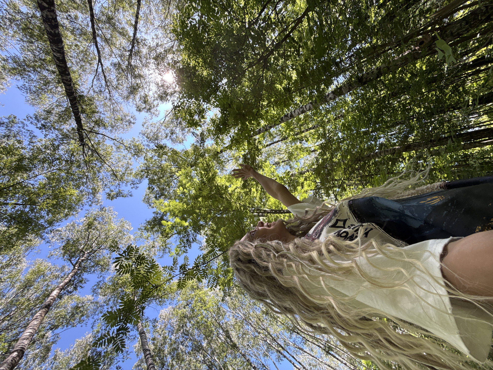 The image captures a vibrant forest scene in Rusanovo, Russia, from a low-angle perspective looking upwards. A fair-skinned individual, likely female, is positioned on the left side, her head turned to the right with a wide smile, gazing skyward. She wears glasses and has long, voluminous blonde hair styled in pronounced waves or dreadlocks that extend past her shoulders. She is dressed in a light-colored, possibly off-white, short-sleeved top with a visible graphic on the right chest area, and a darker, patterned garment underneath. Her right arm is extended upwards and slightly forward, with an open hand, as if reaching towards the sky or foliage. The background is dominated by a dense canopy of deciduous trees, primarily displaying bright green leaves and slender trunks, some with light bark suggesting birch trees. A clear, brilliant blue sky is visible through gaps in the leaves, with a distinct sunburst or lens flare in the upper right quadrant, indicating strong direct sunlight.