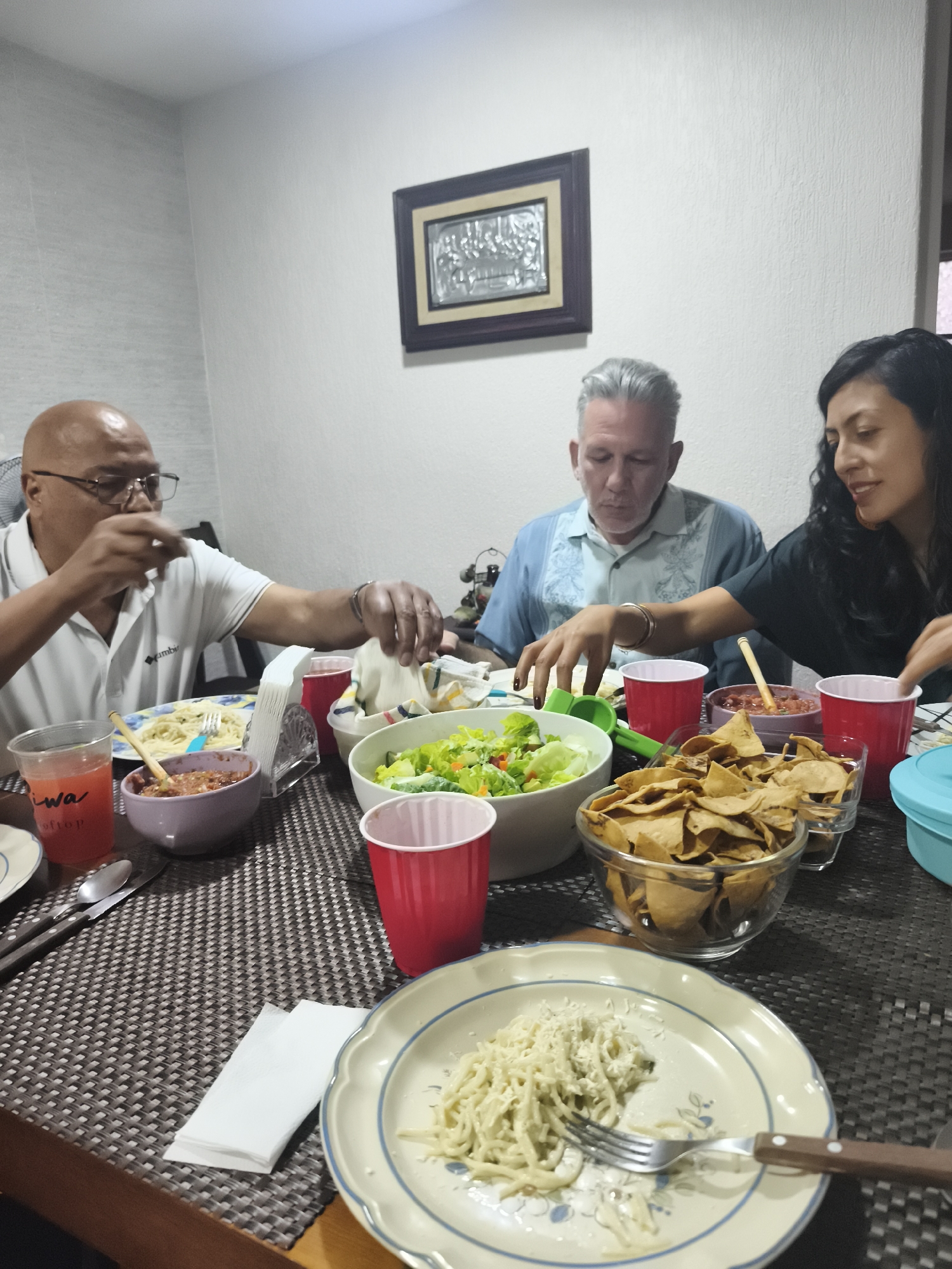Three individuals are seated around a table laden with food. On the left is an African American male wearing glasses and a white shirt with Columbia branding, seemingly in the act of eating. To his right is a Caucasian male with gray hair, dressed in a light blue shirt, also reaching for food. On the far right is a female with long dark hair, wearing a dark top, similarly reaching for the food.
The table is covered with a dark woven mat and holds various dishes, including bowls of spaghetti, salad, salsa, and a bowl of tortilla chips. Several red plastic cups are also present. There are a few plates with food.
In the background, there is a framed picture hanging on the plain wall.
Based on the food and general atmosphere, the location could potentially be a home in Mexico City, Mexico, but there is no definitive evidence within the image to confirm this.