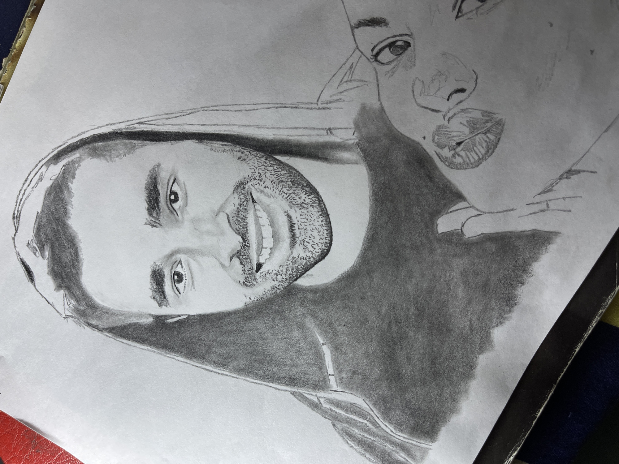 The image displays a graphite pencil drawing on white paper, depicting two subjects. The primary subject is a male figure, likely a young adult, positioned centrally, with short dark hair visible under a dark hooded garment. He has a full beard and mustache, a broad smile revealing teeth, and dark eyes looking directly forward. Below and to the right, a second, less detailed sketch shows features of another person, possibly female, including an eye, part of a nose, and prominent lips, with her head tilted. The drawing exhibits varying degrees of pencil shading, with the male figure being more rendered than the secondary sketch. The white paper itself has slight creases along its lower edge and rests on a multi-colored surface, which includes a dark border and a segment of a red object visible at the top-left. This scene is located in Jos, Nigeria.