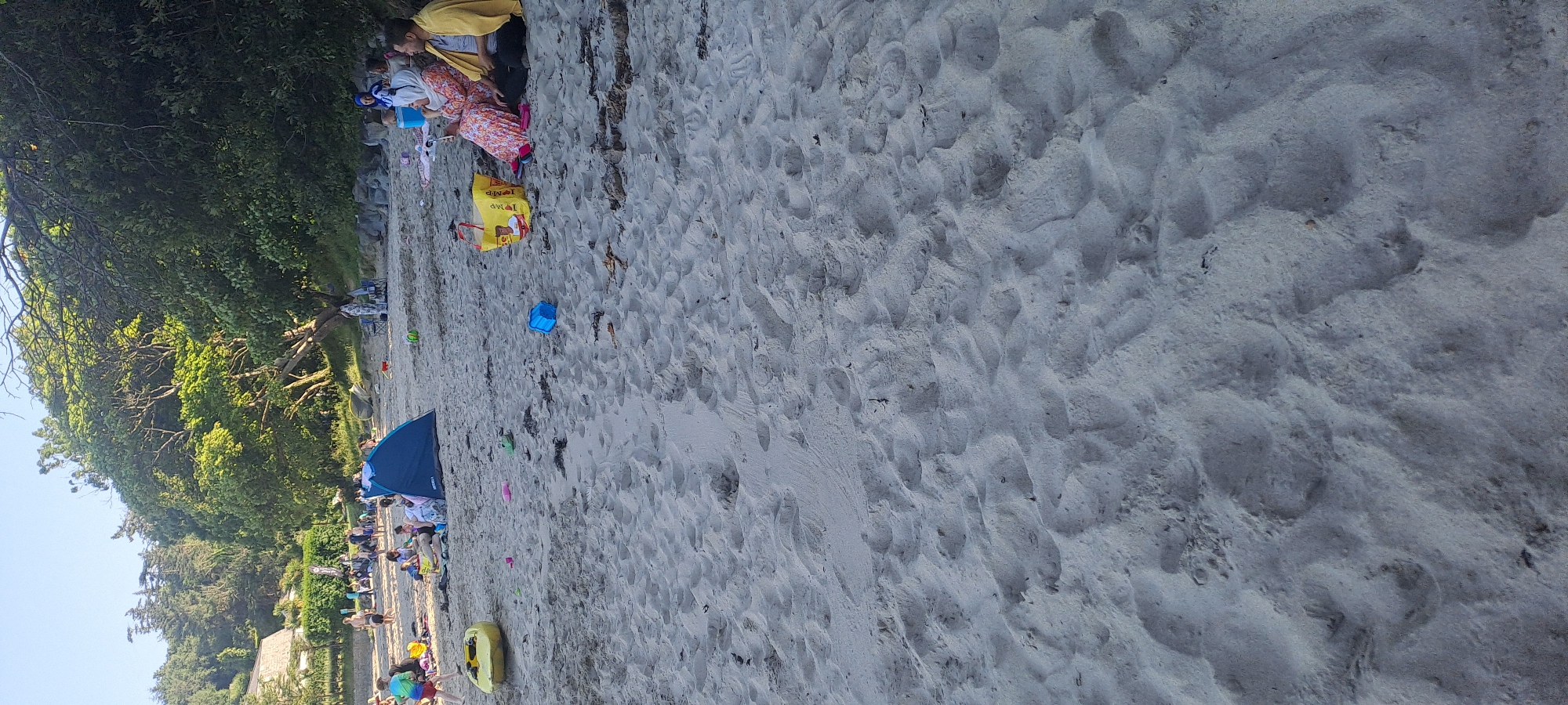 The image captures a sunny day at a beach in an unknown location in Ireland. The foreground is dominated by sand with footprints and some scattered debris. In the midground, a blue tent is pitched on the sand, with several people gathered around it, some sitting and others standing. To the right of the tent, a yellow bag is visible, and further to the right, a couple is seated on the sand, the man wearing a yellow jacket and the woman in a patterned dress and hijab. Behind the beachgoers, a dense line of trees forms the background, with a building visible through the trees on the far left. Various beach toys, including a yellow kayak-like object, and buckets are scattered across the sand. The overall scene depicts a relaxed day at the beach with families enjoying the outdoors.