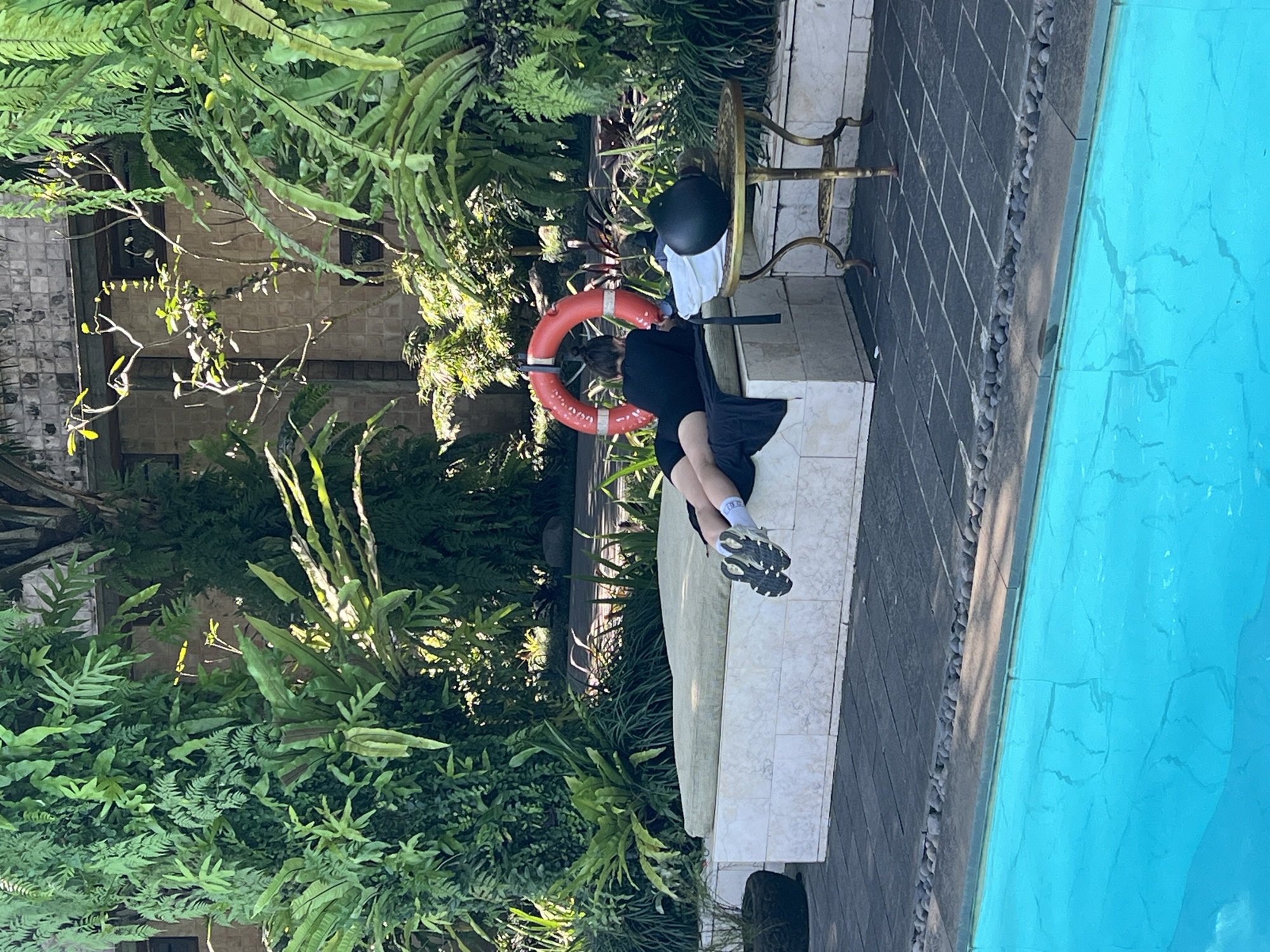 The image depicts a person sitting on a tiled bench next to a swimming pool. The pool's water is bright blue. Dense green vegetation surrounds the area. A life preserver is propped against the back of the bench. A small table with a black helmet on it sits nearby. The scene is located in Petulu, Indonesia.