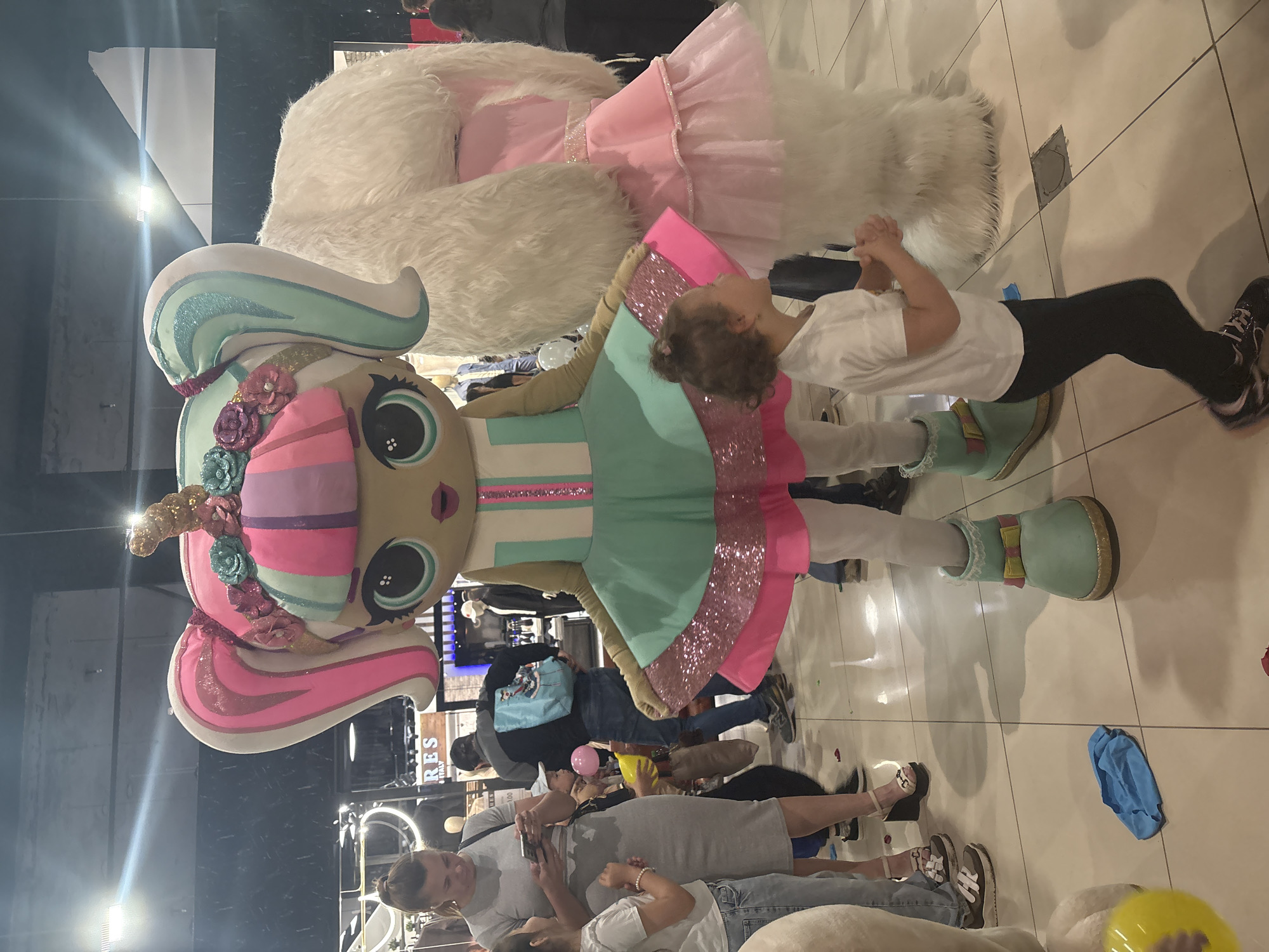 Captured indoors, the scene shows a child interacting with a large doll-like character wearing a multicolored dress and shoes. The doll has large eyes and hair with vibrant pink and blue. The child, dressed in a white shirt and black leggings, stands close to the doll with clasped hands. Numerous people are visible in the background, some of whom are looking at phones or cameras, likely documenting the scene. A fluffy white animal costume is partially visible to the right. The flooring is a light-colored tile. In the background, elements such as clothing racks and a shop sign can be seen. The location appears to be a shopping mall or retail environment, possibly in the city of Tri Protoka, Russia.