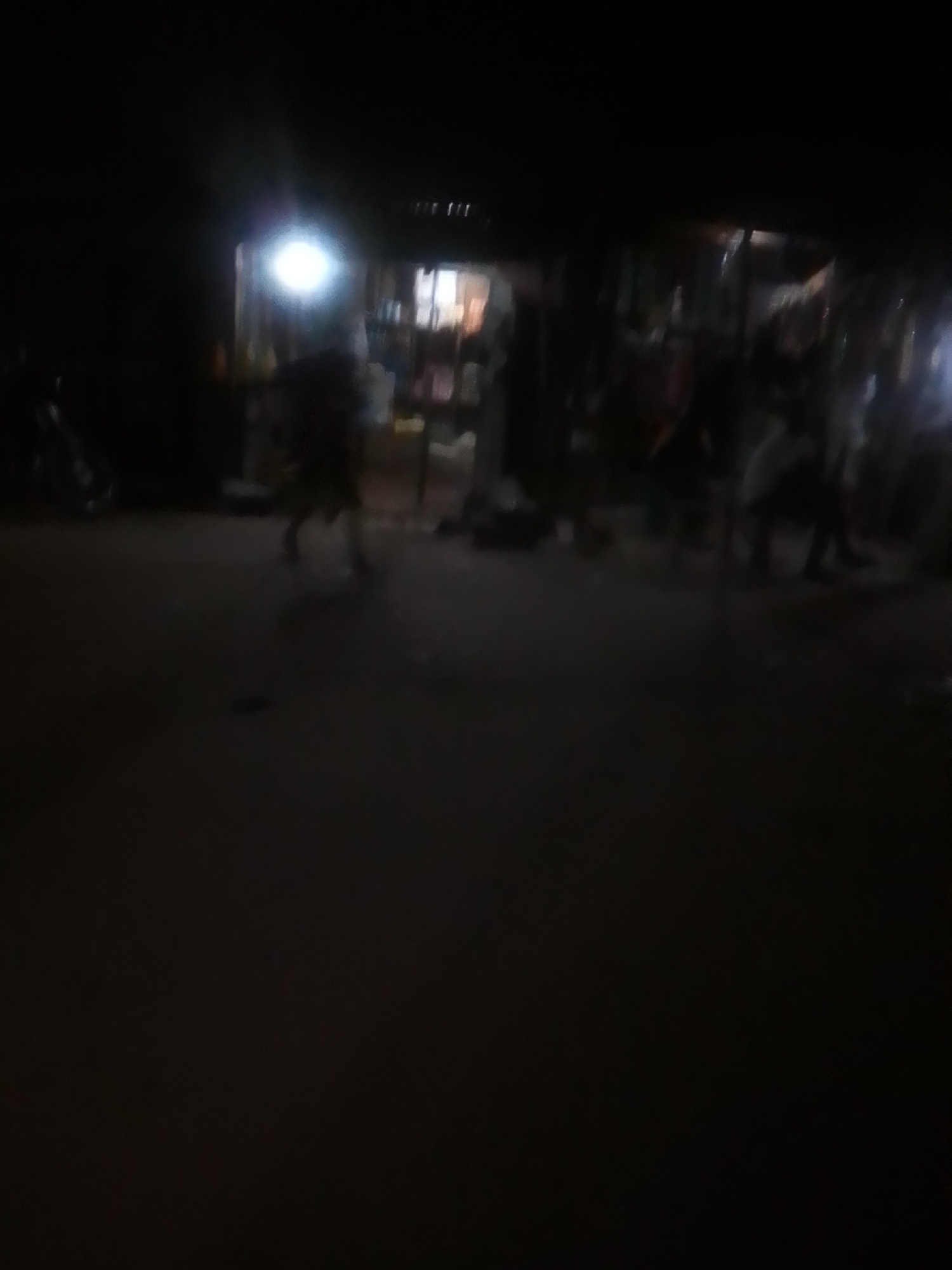 The image depicts a dimly lit street scene in Kwadon, Nigeria. In the background, there appears to be a market or shop area with several individuals. A bright light emanates from within this area, casting some illumination on the surroundings. The foreground is largely dark, with some indistinct shapes that could be people or objects on the ground. The overall impression is one of a busy, albeit poorly lit, evening or night scene. The resolution is low, making it difficult to discern fine details of the people or the environment.