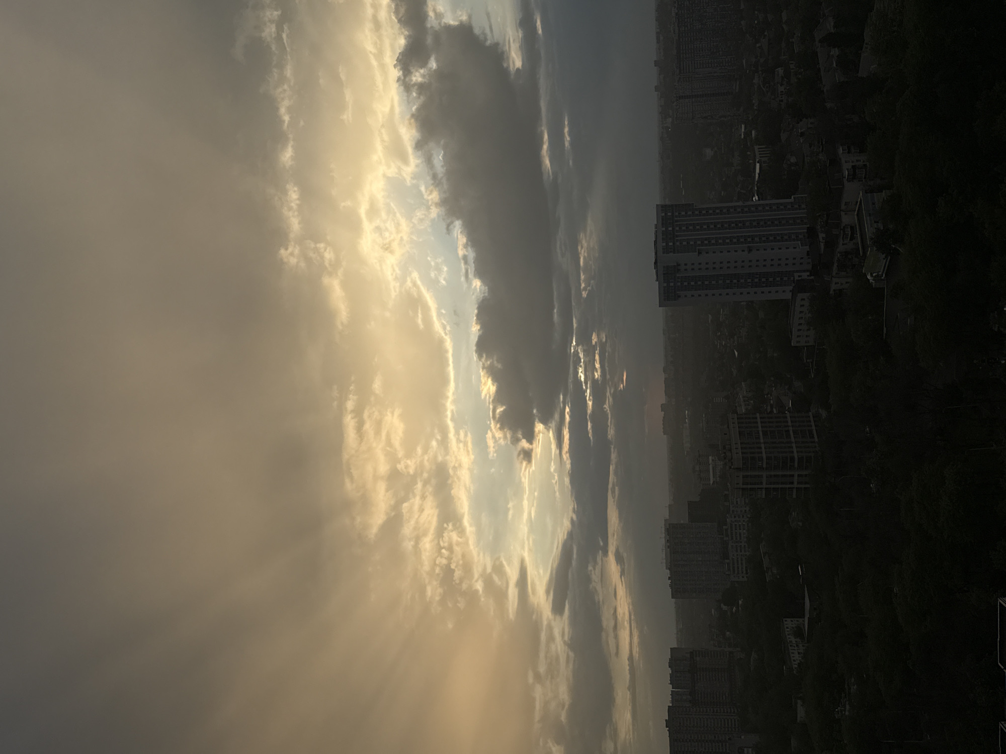 The image depicts a cityscape in Rostov-on-Don, Russia, under a dramatic sky. The right side of the frame shows buildings, some high-rise, amidst trees. The sky is filled with clouds, with the sun filtering through, creating rays of light. The overall lighting suggests either sunrise or sunset. The general scene is an urban environment in the midst of atmospheric weather conditions.