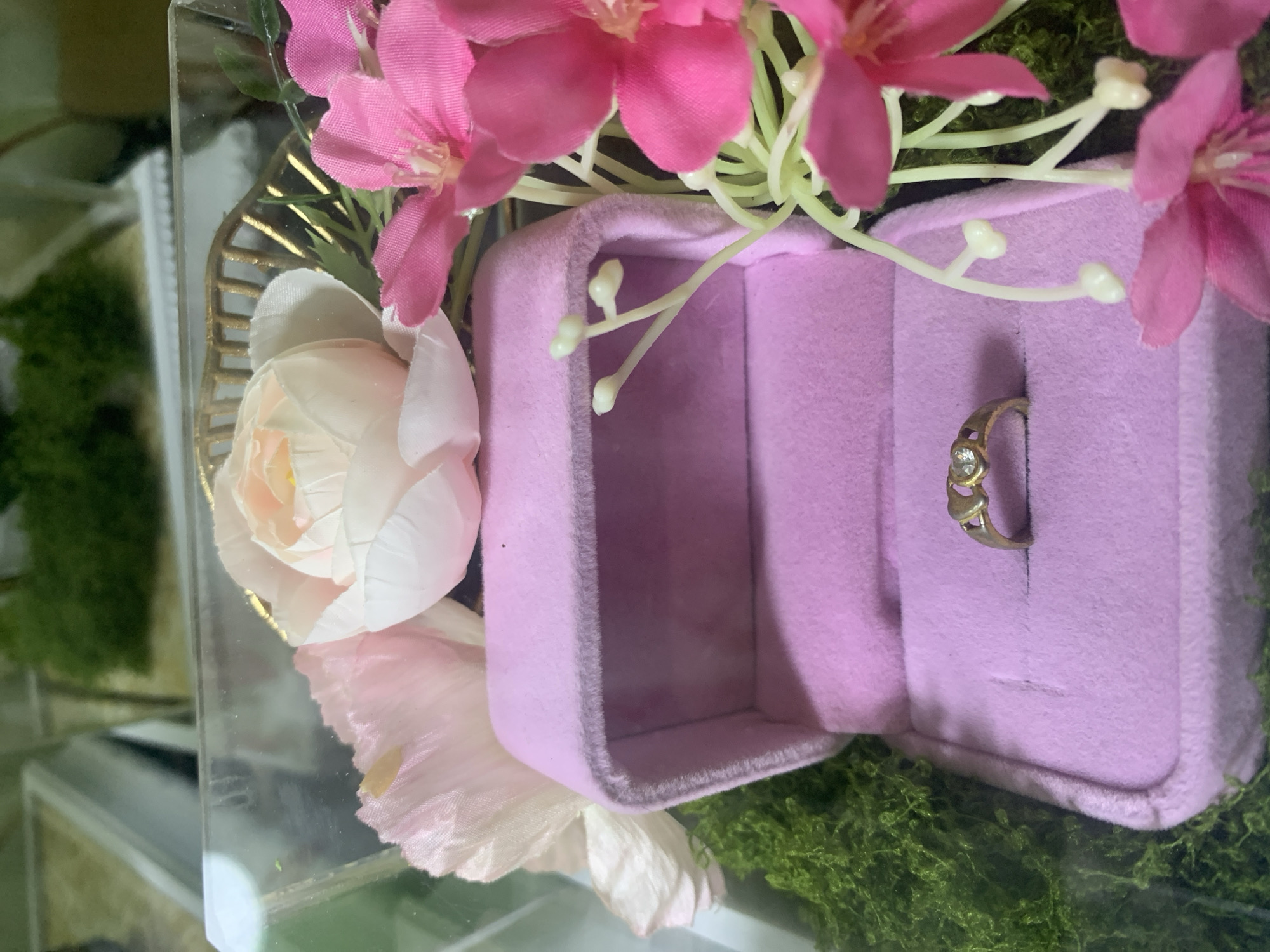 A gold ring with a heart-shaped design and a small diamond is displayed inside a pink velvet ring box. The box is part of a larger decorative arrangement that includes artificial flowers in shades of pink and white, and green faux moss. The arrangement is enclosed in a clear plastic box, creating a diorama-like presentation. The background is blurred, showing hints of greenery and reflective surfaces. The scene appears to be a product display or a gift presentation. The location in Kuala Pembuang, Indonesia, is not visually evident from the provided image.