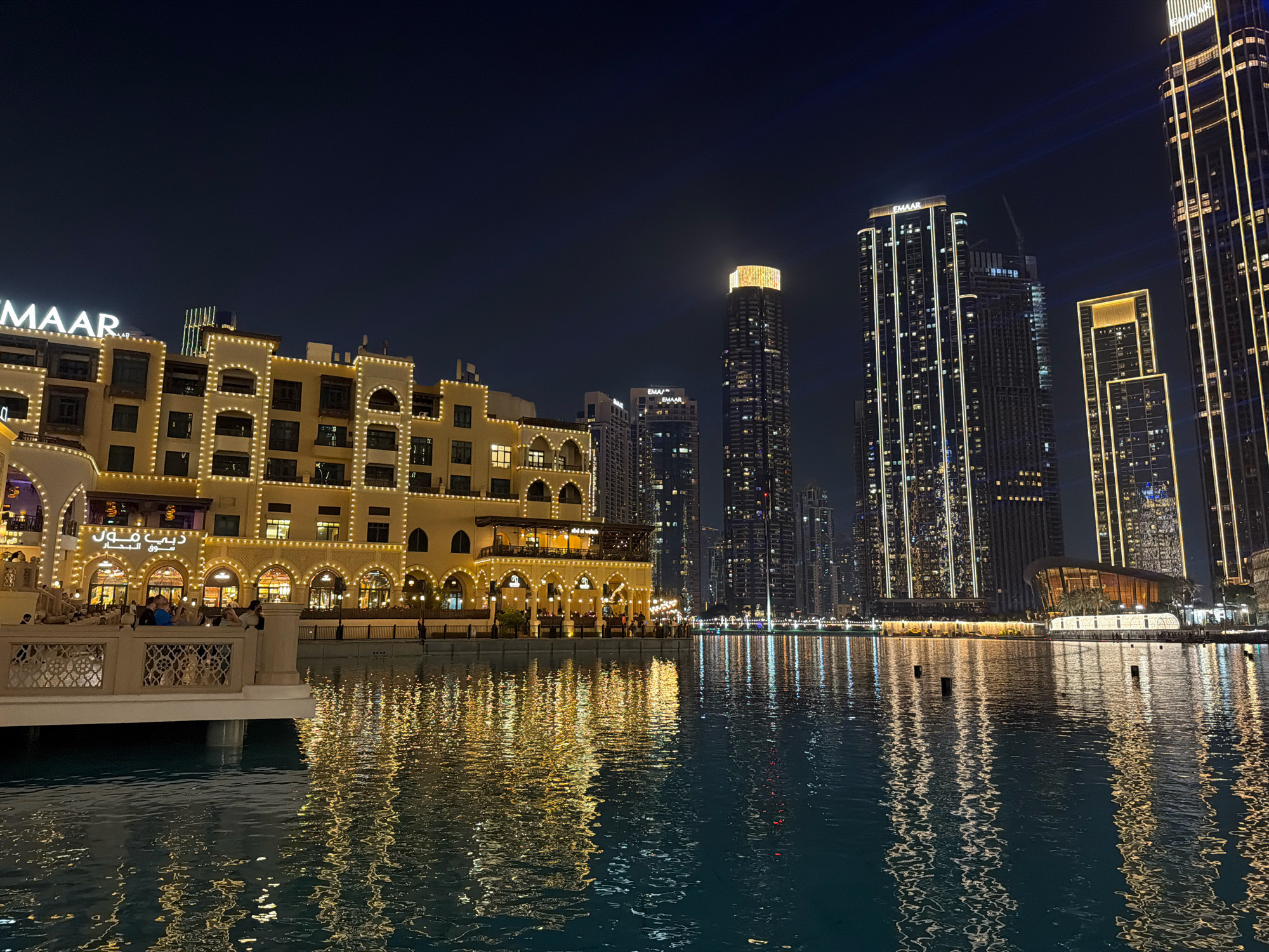 The image depicts a nighttime scene in Dubai, United Arab Emirates, likely at the Dubai Fountain or a similar waterfront area adjacent to the Burj Khalifa and Dubai Mall. In the foreground, a body of water reflects the warm, golden lights of surrounding buildings. On the left, a low-rise building with arched windows and decorative lighting is visible, displaying Arabic script and the "EMAAR" logo. Several people are seen on a balcony or walkway in front of this building. In the background, several towering skyscrapers, illuminated with vertical lines of light, dominate the skyline, characteristic of Dubai's modern architecture. One prominent skyscraper features a brightly lit, golden crown. The dark night sky provides a stark contrast to the illuminated structures. The scene conveys a sense of urban vibrancy and a well-lit cityscape at night.