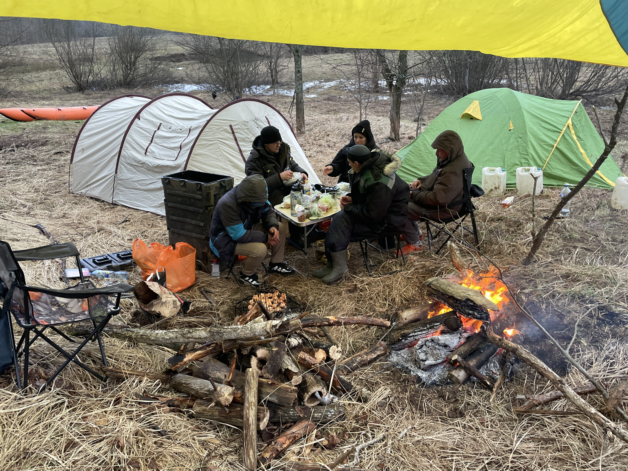 The image depicts a campsite scene, likely in a natural environment near Zabor'e, Russia. The foreground shows a campfire with burning logs and a pile of additional firewood. A grill sits over some coals, possibly for cooking food. In the center, five people are gathered around a small table, which is covered with food, drinks, and various containers. They appear to be eating or preparing food. To the left, there is a collapsible camping chair and a crate with bags. In the background, two tents are visible: a larger white tent and a smaller green tent. Beyond the tents, there is a sparse wooded area with signs of snow on the ground. An orange inflatable boat also lies to the left. A yellow and blue tarp is suspended above the people and the table.