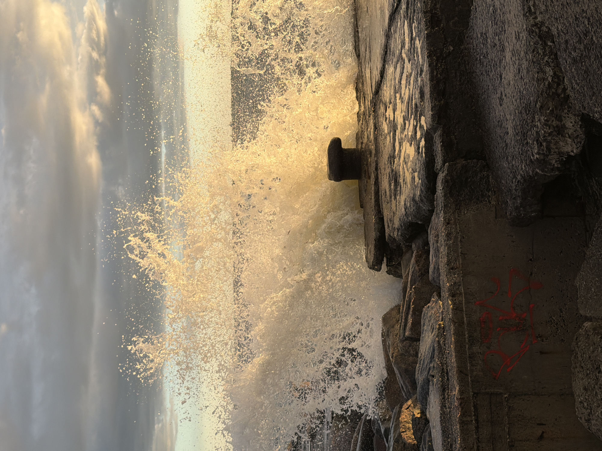 A wave crashes against a rocky shore and concrete pier at sunset in Tallinn, Estonia. The water spray catches the golden light of the setting sun, creating a luminous and dynamic effect. A dark, mushroom-shaped bollard stands on the pier, a weathered post designed to tie up boats. The foreground features rough, gray rocks and concrete structures, some of which are covered in red graffiti. The sky above is a mix of dark, brooding clouds and soft, illuminated patches of light.